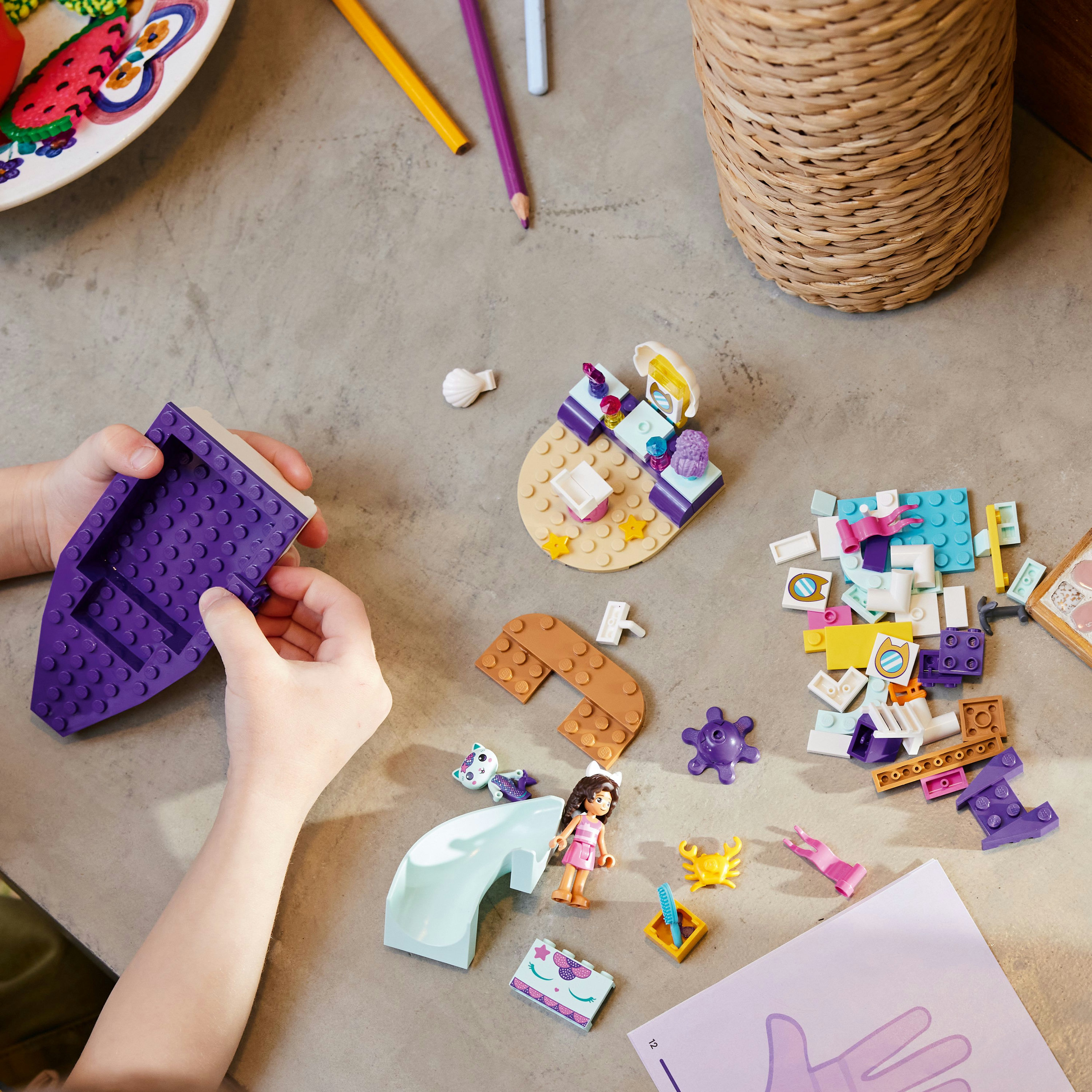 Hände montieren Lego-Steine. Lila Basis, gelber Sand und Figuren. Grauer Tisch.