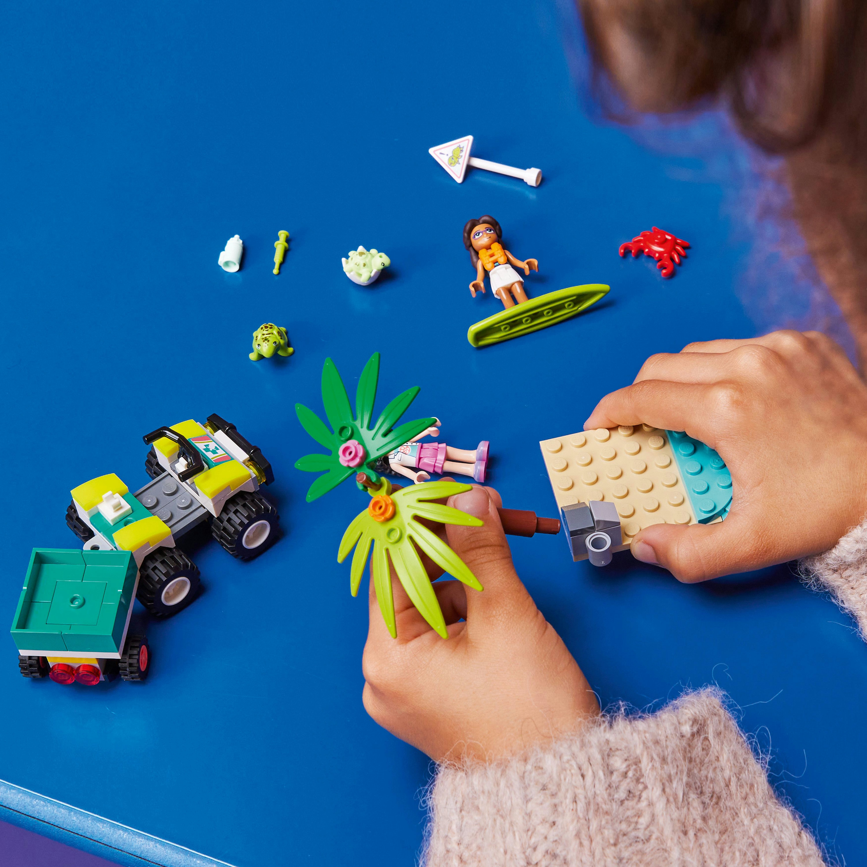 Hände, die ein LEGO-Set auf einer blauen Oberfläche zusammenbauen. Das Set umfasst ein kleines Auto, eine Krabbe und eine Palme.