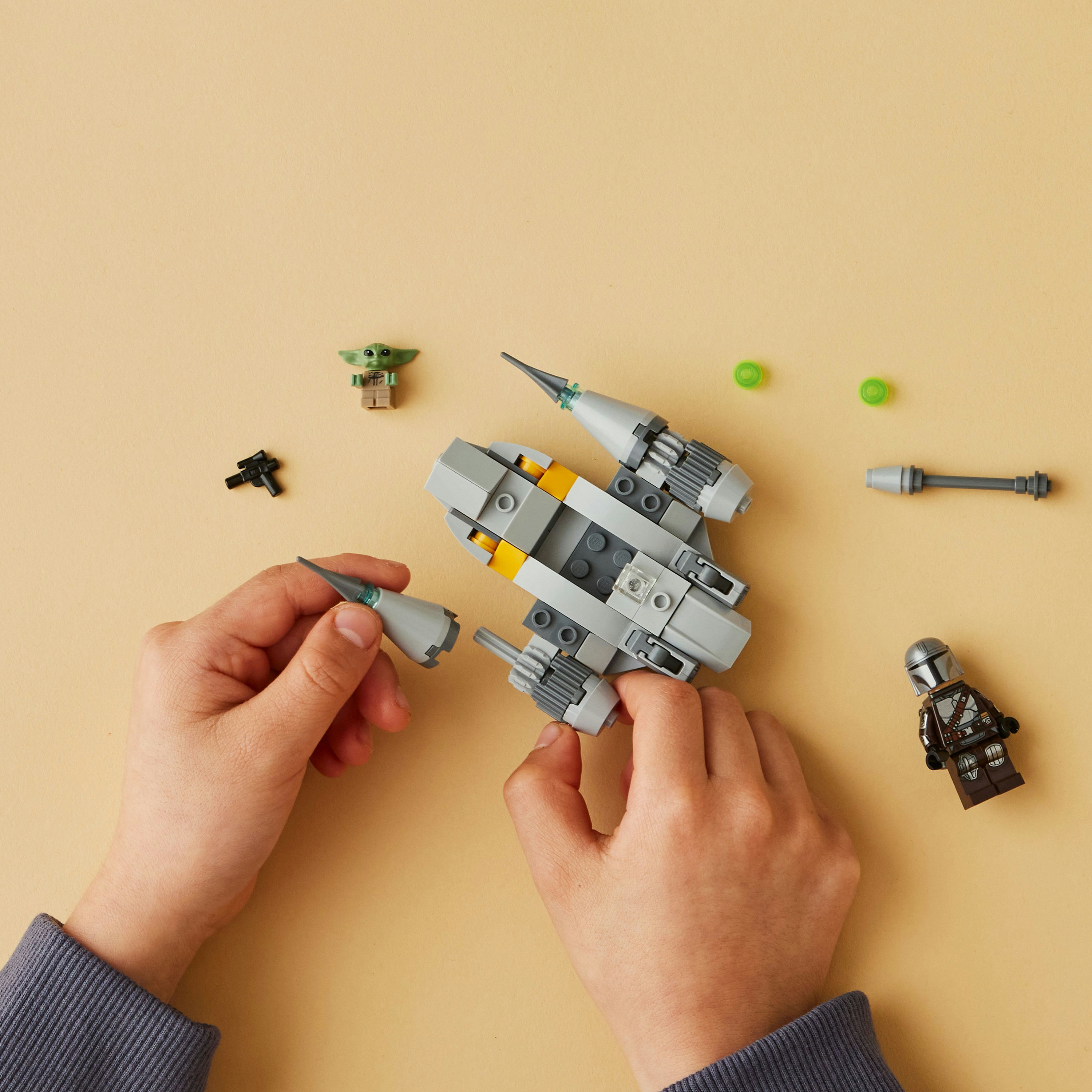 Hände, die ein graues und gelbes Raumschiff mit Grogu, Mandalorian und anderen Lego-Teilen zusammensetzen.