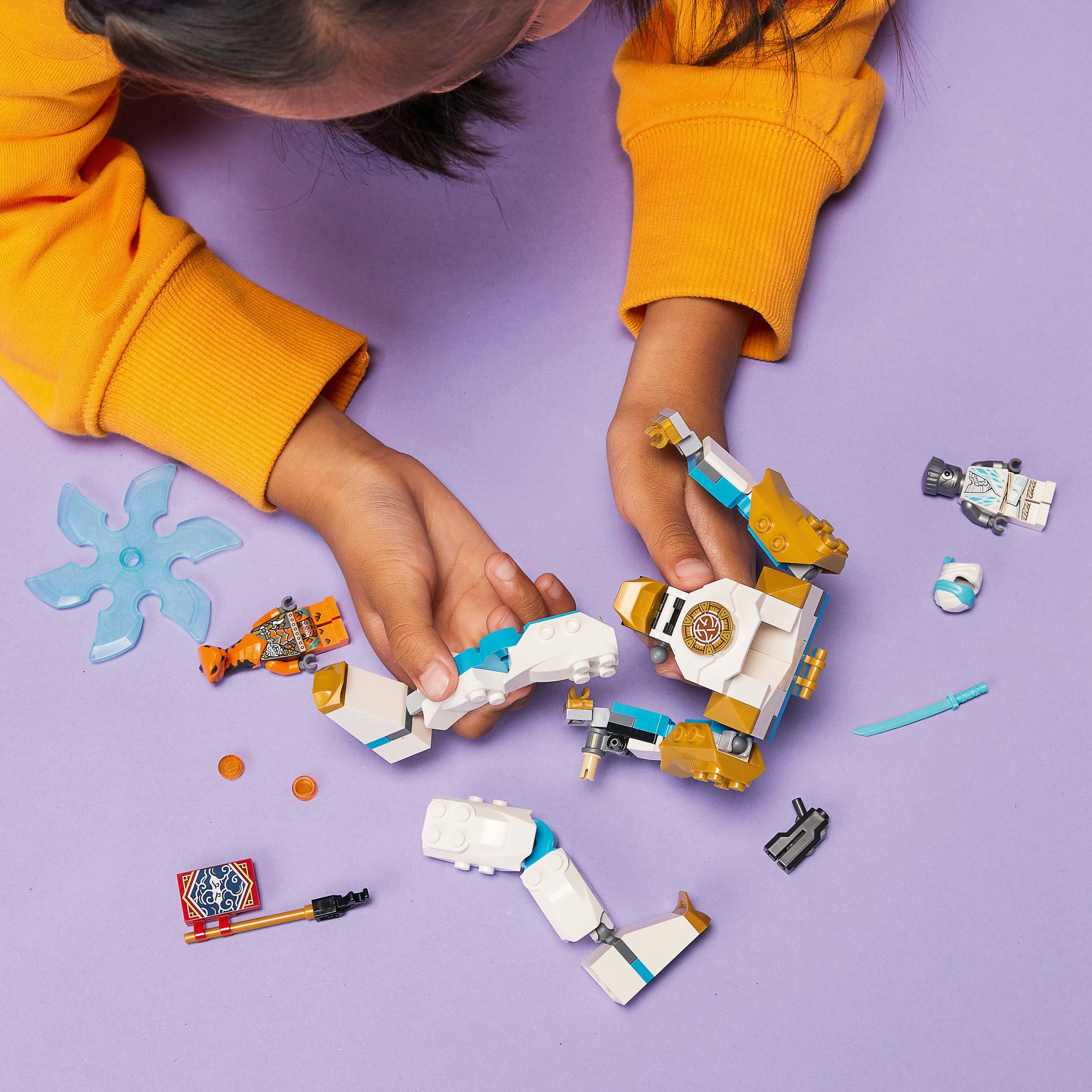 Un bambino gioca con pezzi Lego, tra cui parti di robot, uno shuriken e minifigure, su una superficie viola.