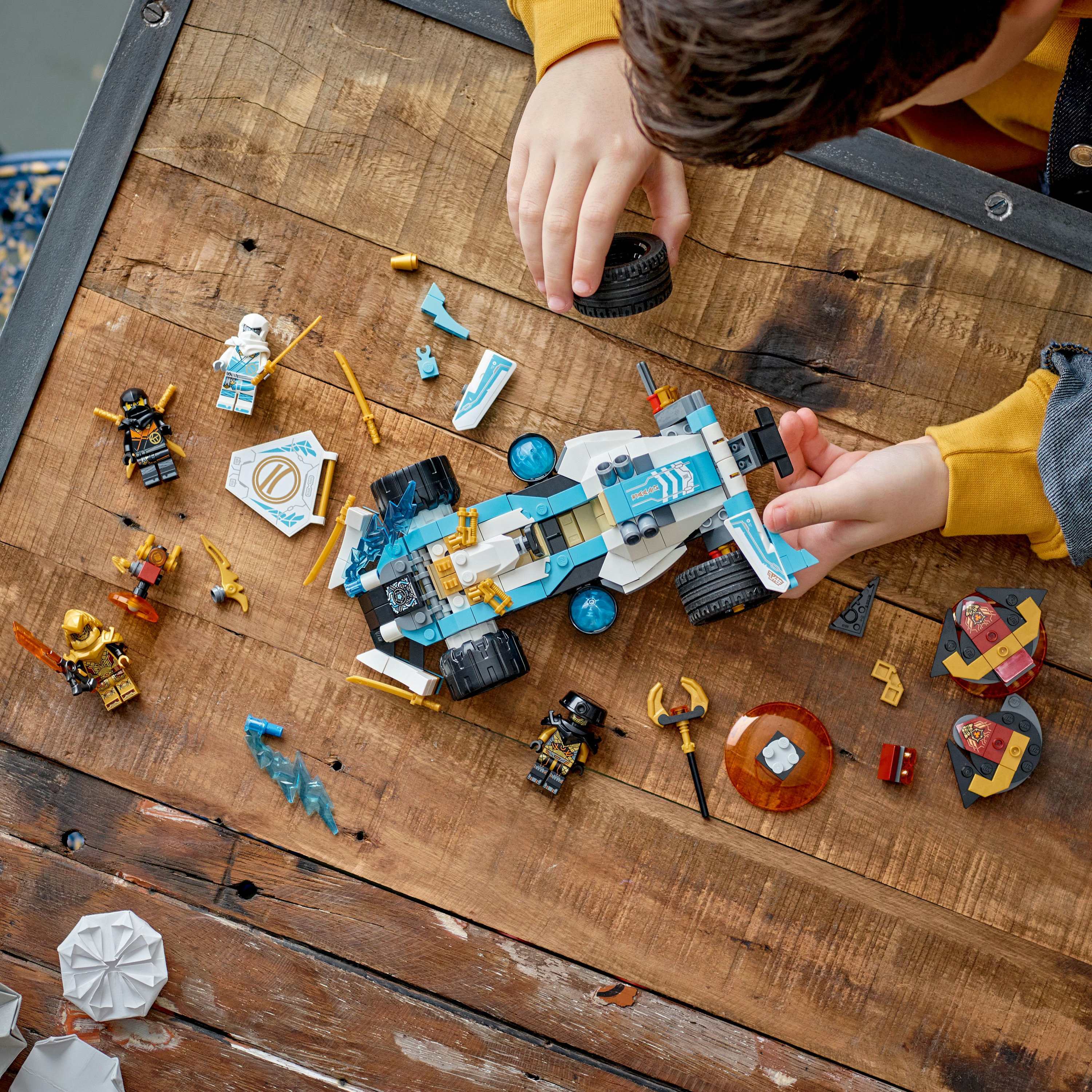 Un enfant joue avec un set Lego Ninjago. Des figurines et des pièces de véhicules sont éparpillées sur une table en bois.