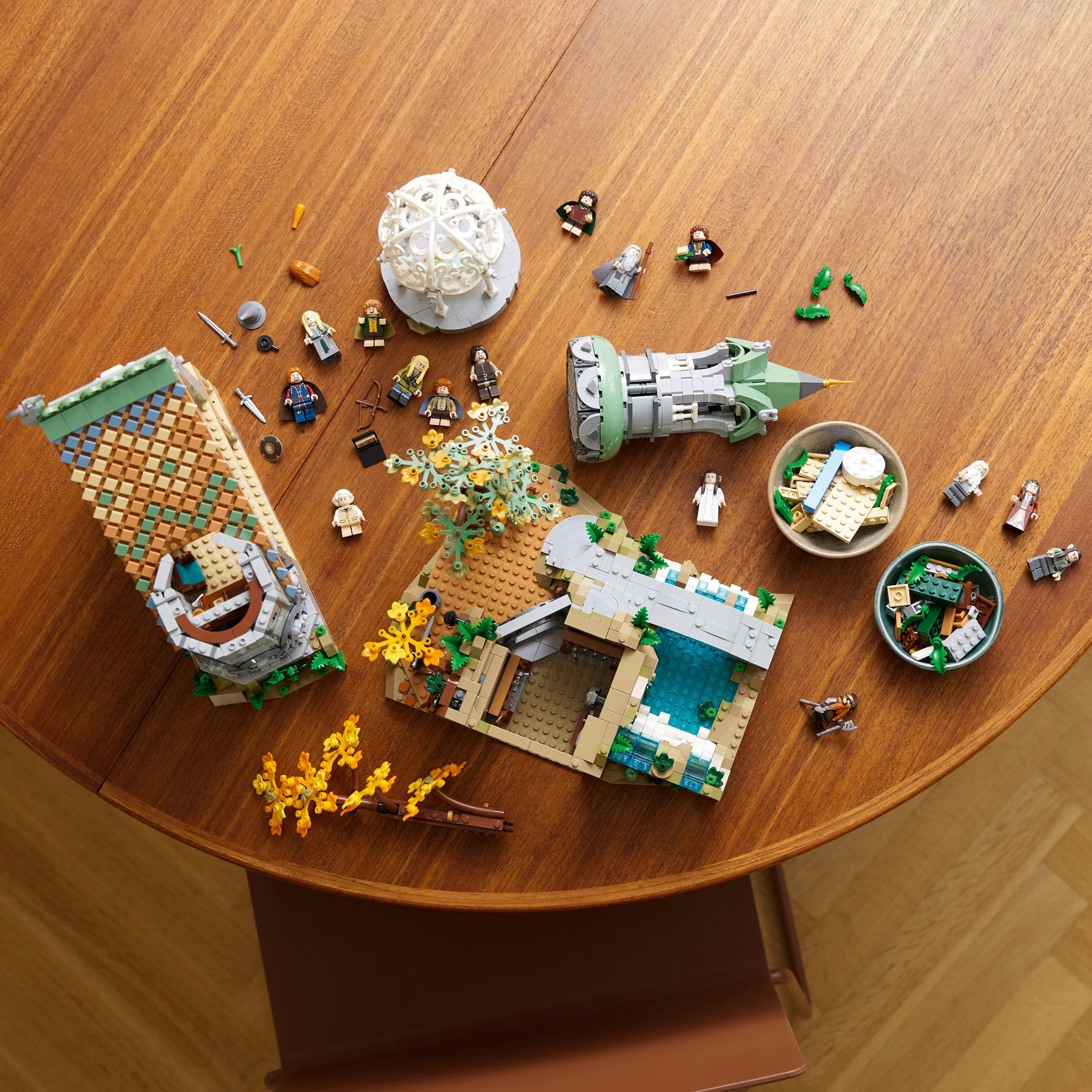 Scena Lego na stole z różnymi minifigurkami i elementami architektonicznymi. Widoczne małe drzewo i basen.