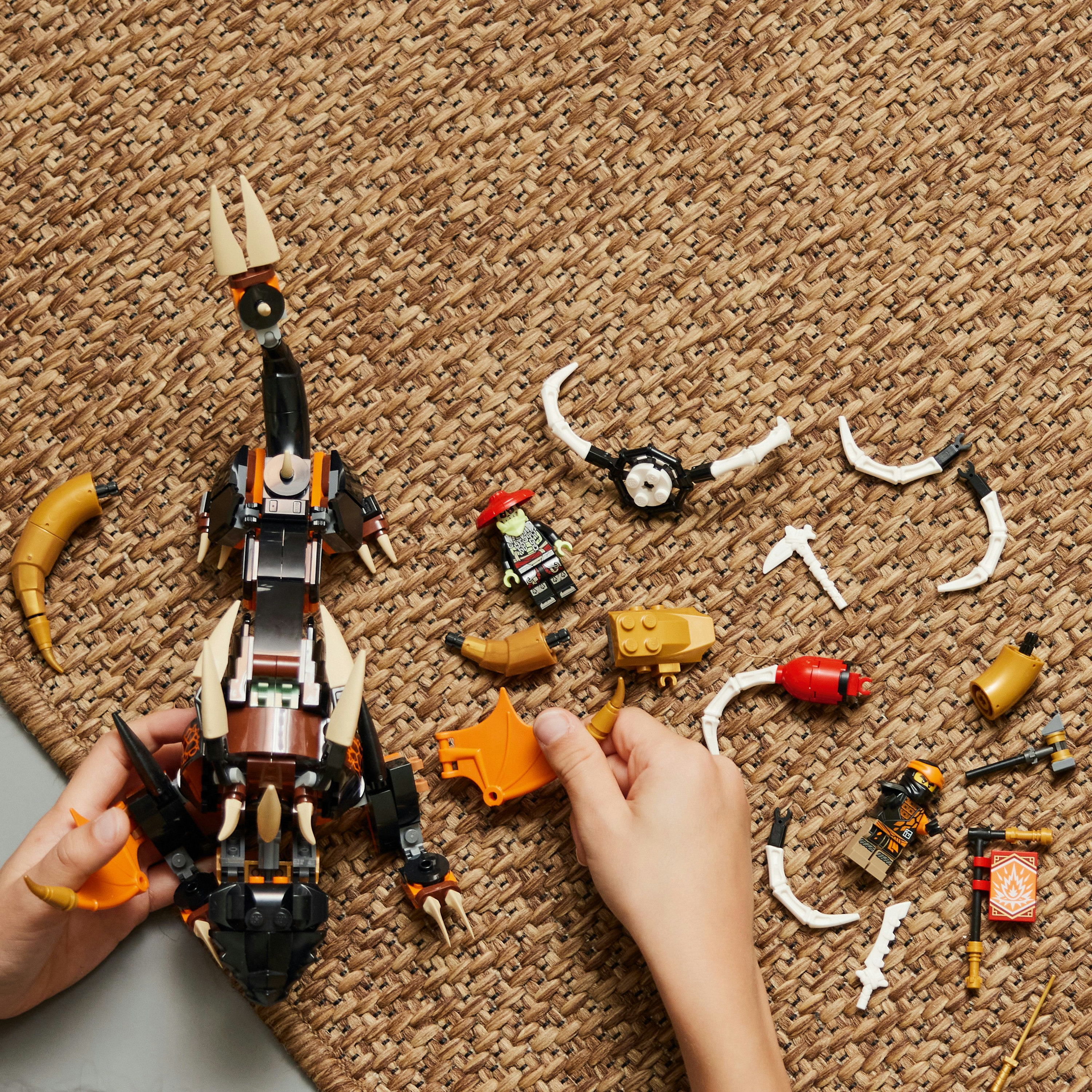 Hände, die eine Lego-Drachenfigur zusammensetzen, mit verschiedenen Teilen, die auf einem Teppich verstreut sind.