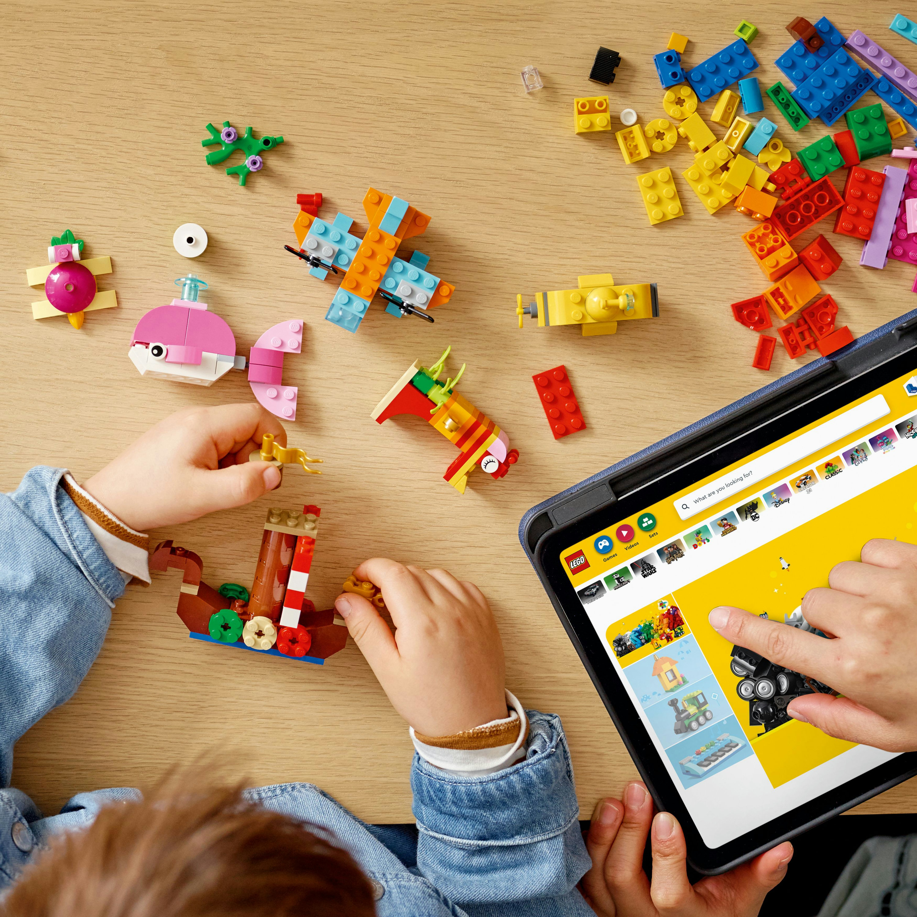 Kinderen bouwen met kleurrijke Lego-stenen op een houten tafel. Een tablet toont een Lego-app.