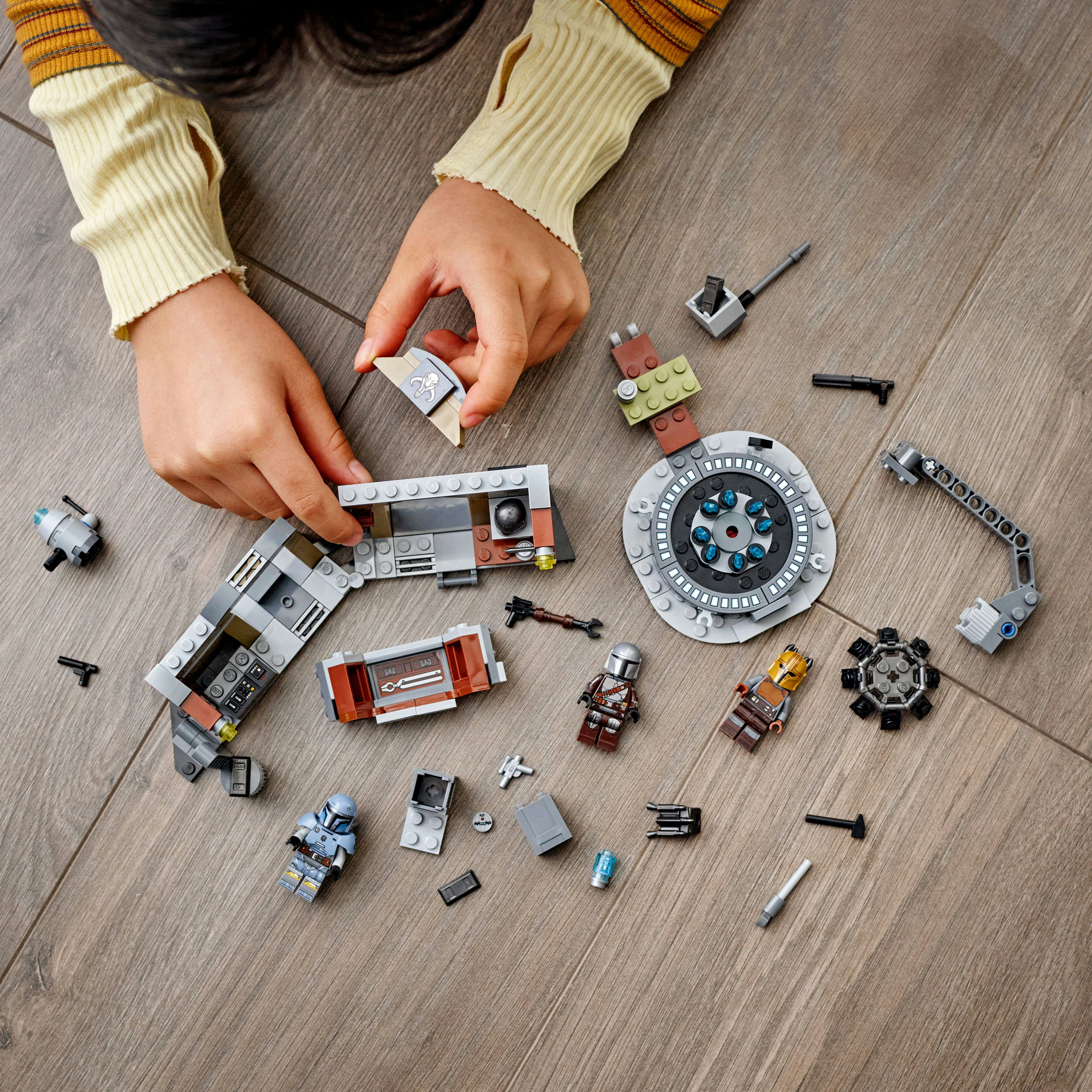 Hände, die Lego-Teile anordnen, mit verschiedenen Figuren und Bauteilen auf dem Boden verstreut.