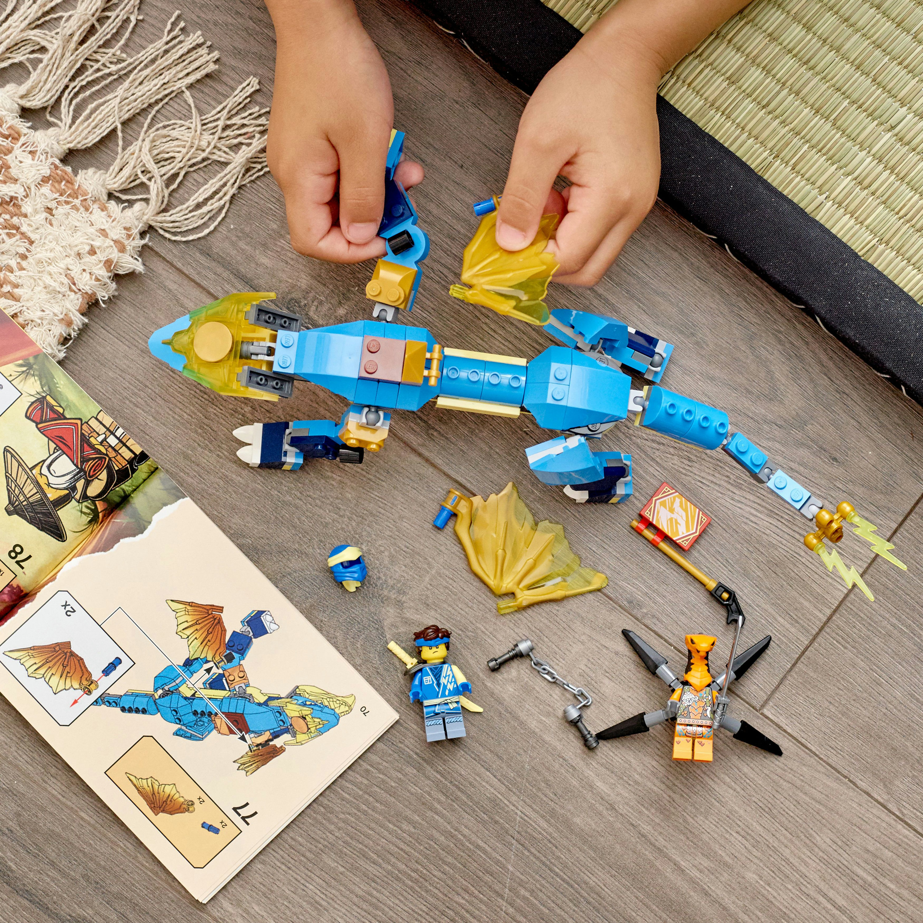 Hände bauen einen blauen Lego-Drachen mit gelben Flügeln und Minifiguren auf Holzoberfläche zusammen.