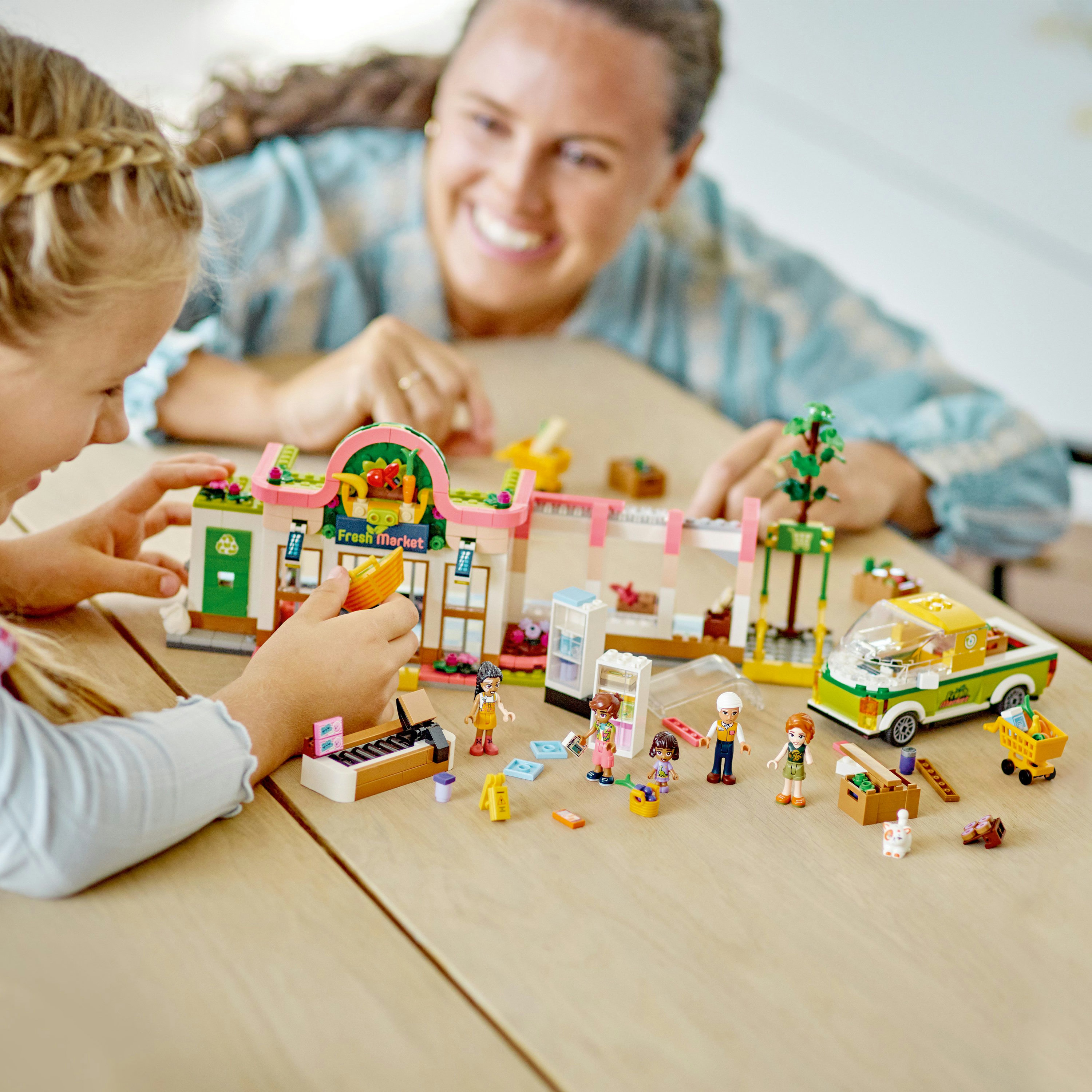 Mutter und Tochter spielen mit einem LEGO-Markt-Set, das Figuren und ein Auto auf einem Holztisch zeigt.