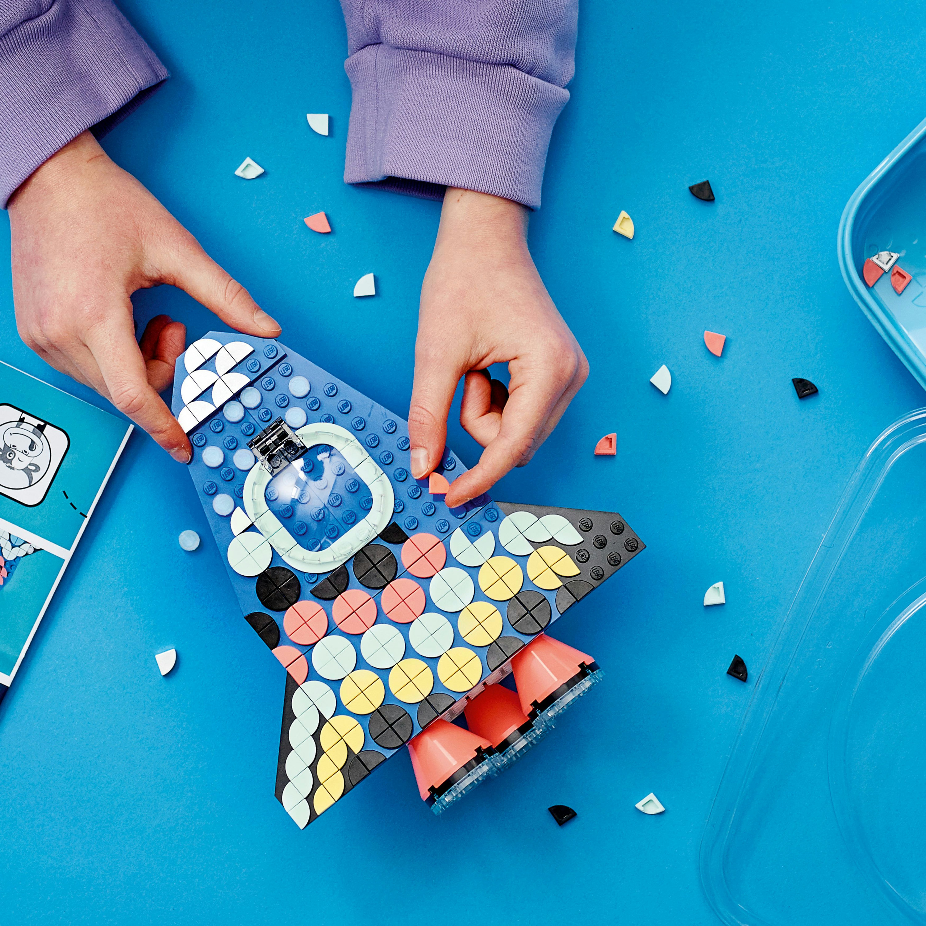 Ręce budujące niebieską rakietę Lego z kolorowymi okrągłymi elementami i innymi elementami Lego.