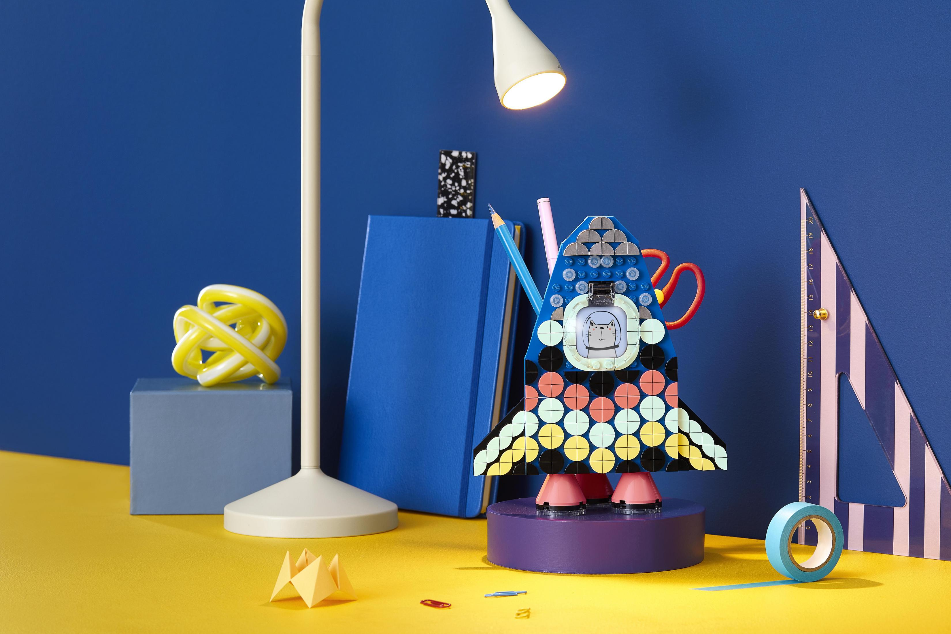 Kolorowa rakieta Lego na biurku z lampą, książką i innymi przedmiotami.