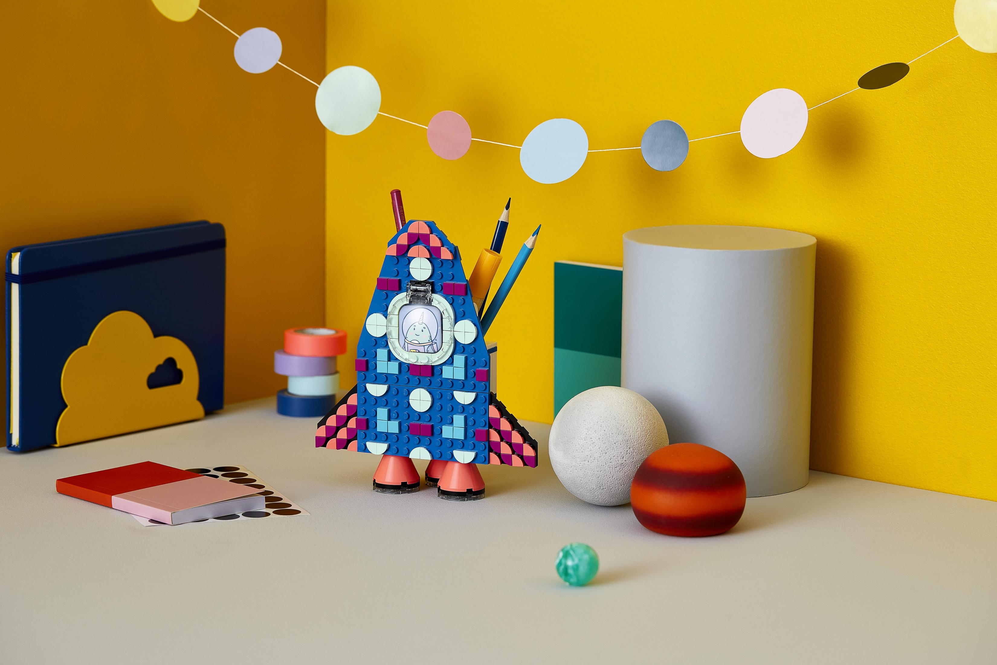 Kolorowy uchwyt na ołówki w kształcie rakiety LEGO z długopisami i ołówkami, otoczony elementami dekoracyjnymi.