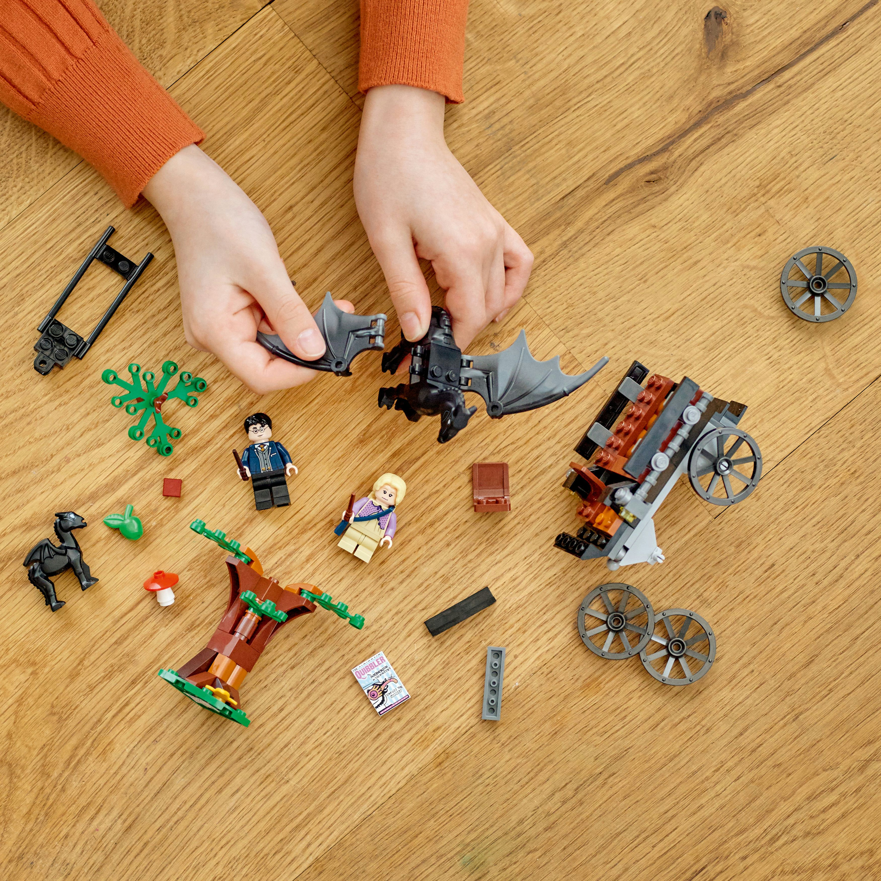 Mani che assemblano una scena Lego con vari elementi: un carro, figure, un albero e piccoli pezzi su legno.