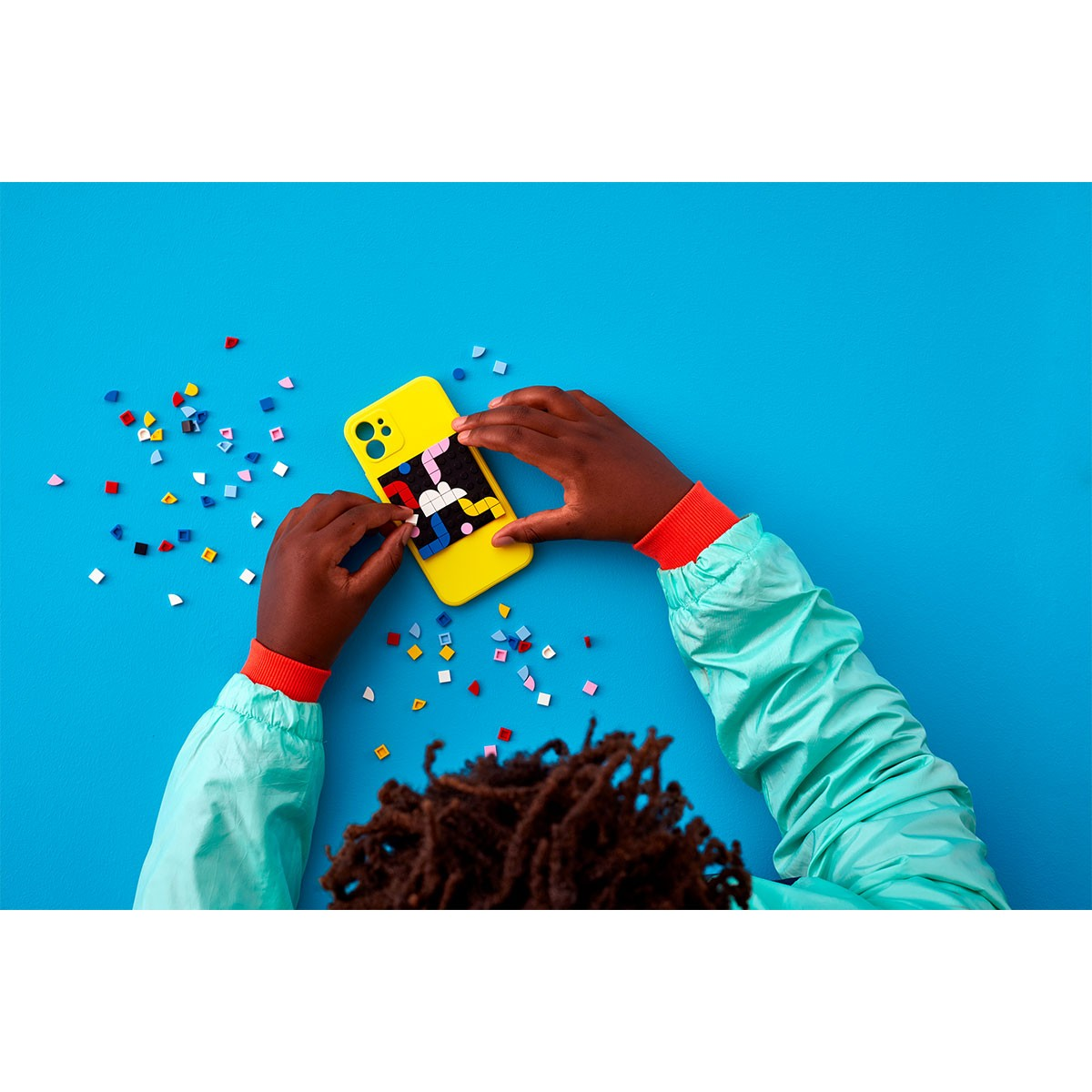 Mani che tengono una custodia per telefono gialla con arte Lego colorata su uno sfondo blu cosparso di pezzi Lego.