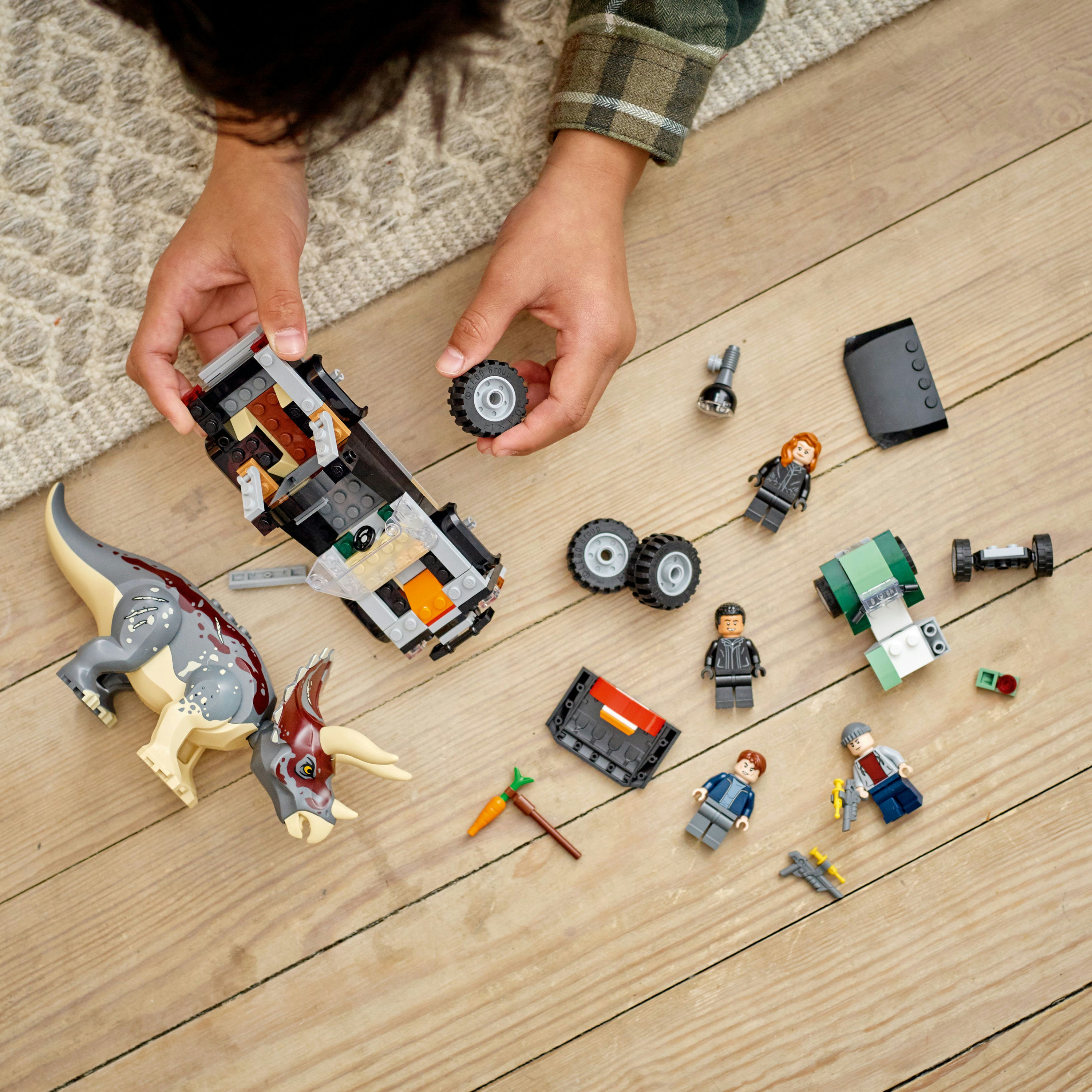 Set Lego con auto, dinosauro e personaggi, sparsi su pavimento di legno, mani di bambino visibili.