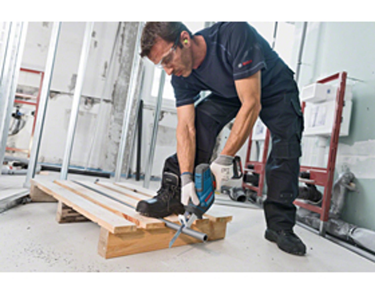 Mężczyzna tnie rurę narzędziem, stojąc na drewnianej palecie na placu budowy.