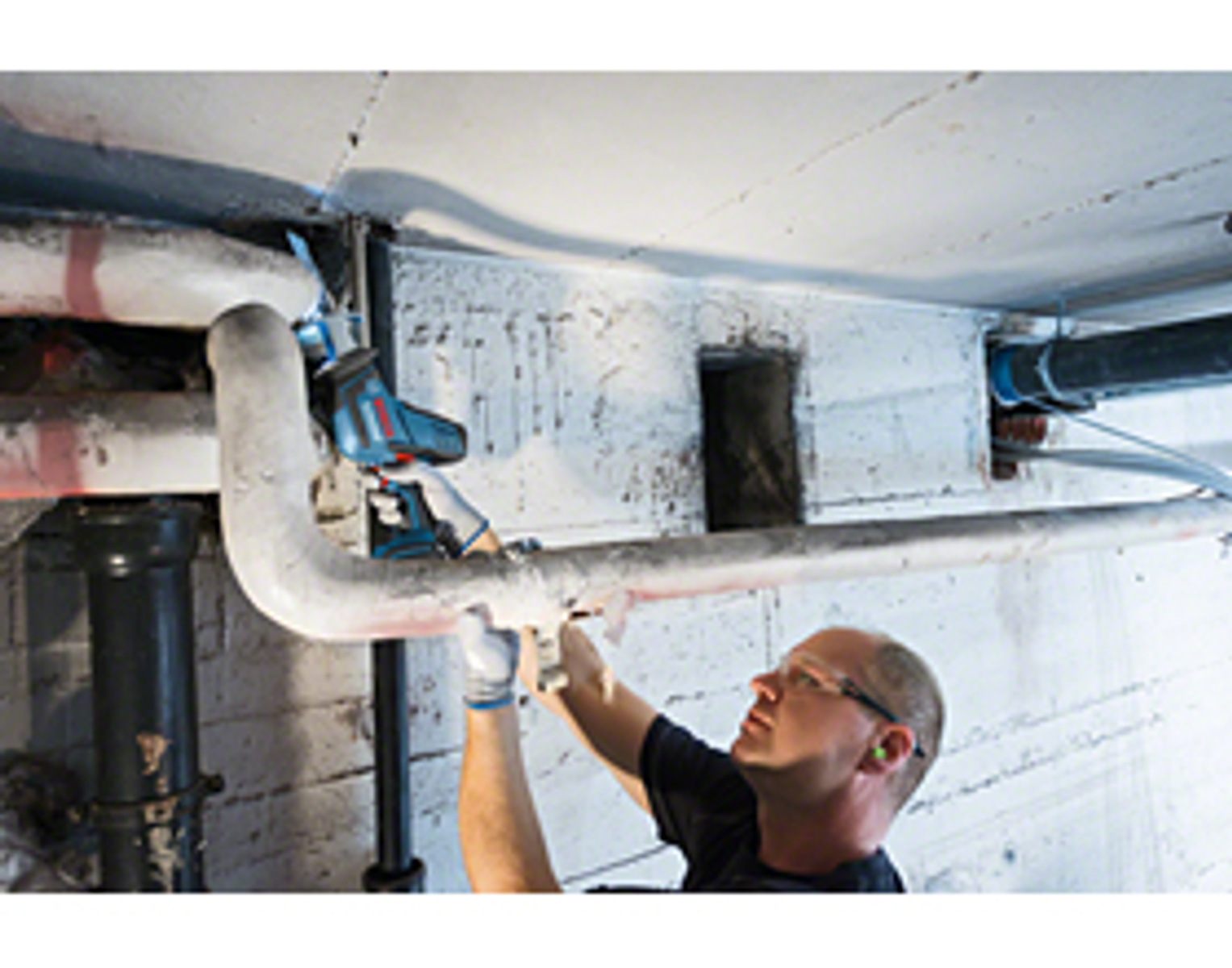 Mężczyzna tnący rurę elektronarzędziem, w okularach ochronnych, w betonowym pomieszczeniu.