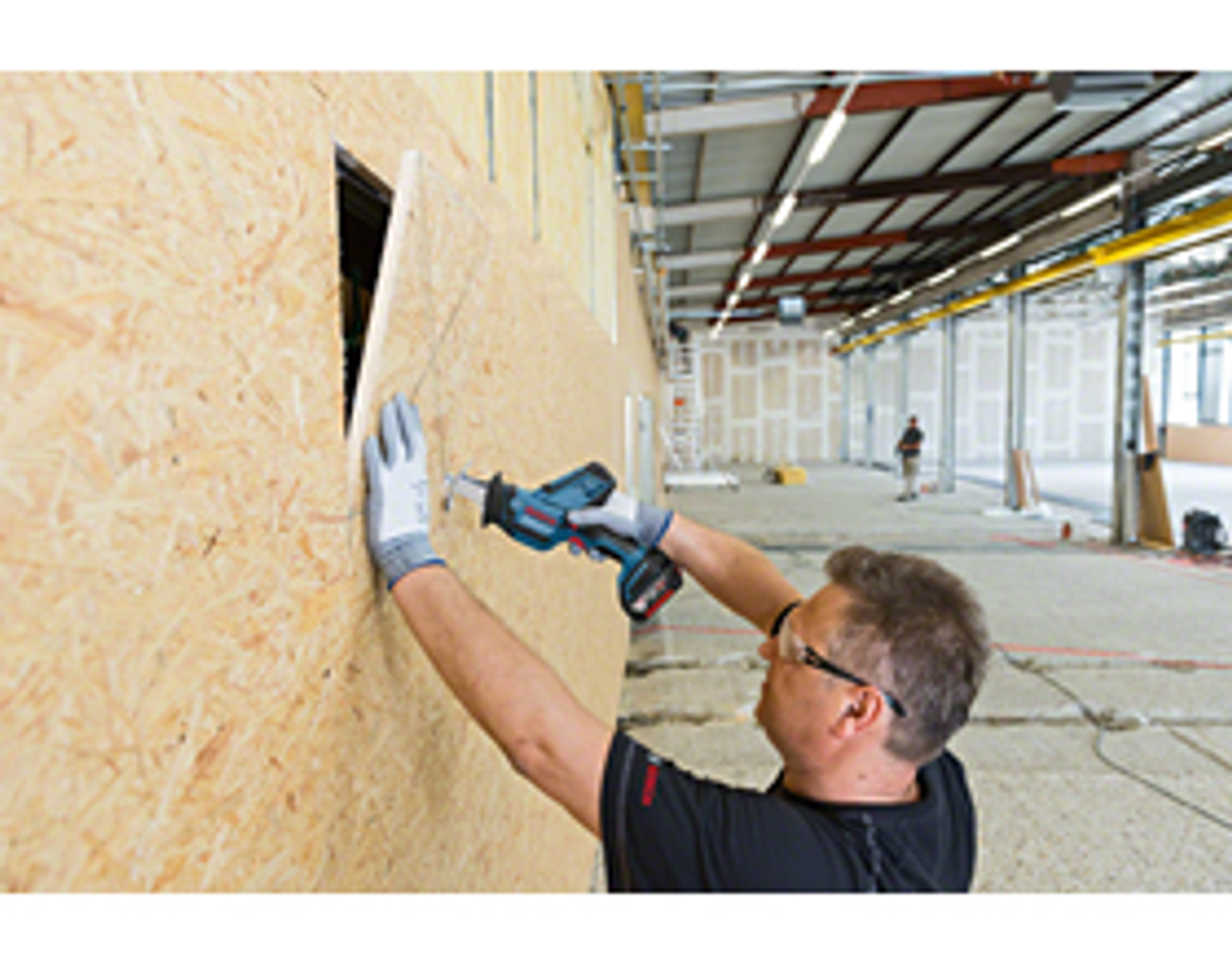 Mężczyzna używający piły do cięcia ściany. Ściana jest wykonana z drewna. Tło to plac budowy.