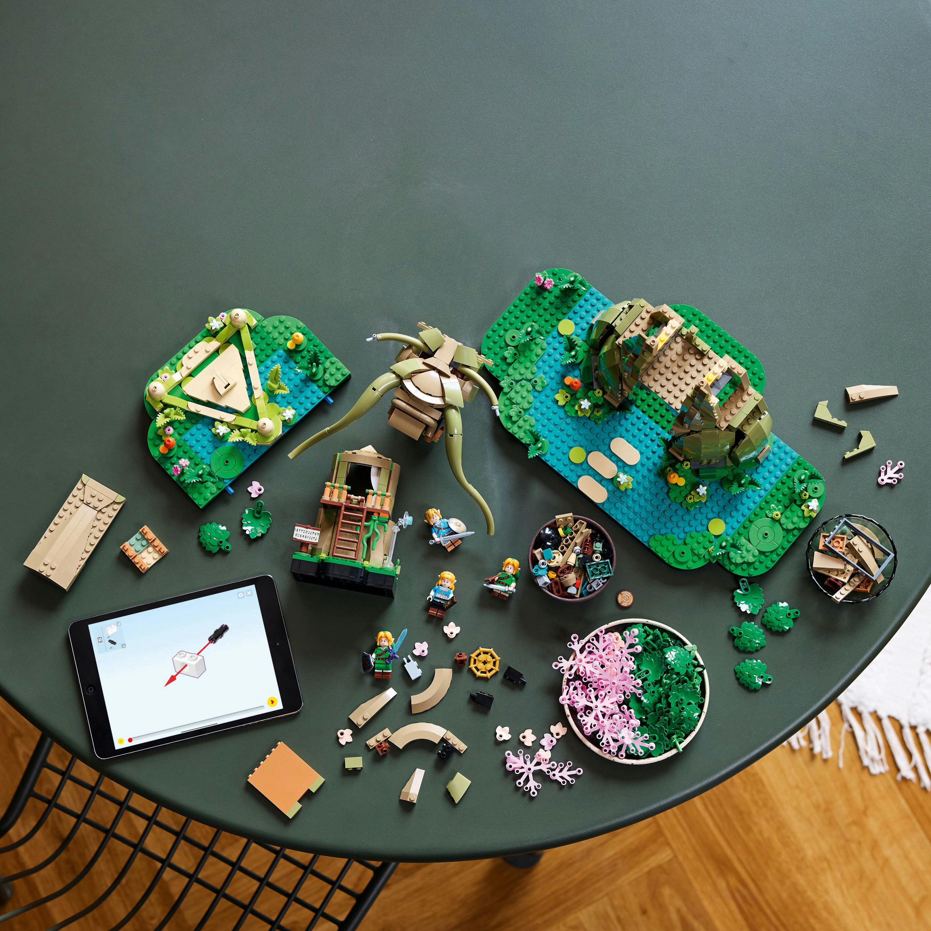 Elementy zestawu Lego na zielonym stole. Zawiera złożone sceny, luźne elementy i tablet z instrukcjami.
