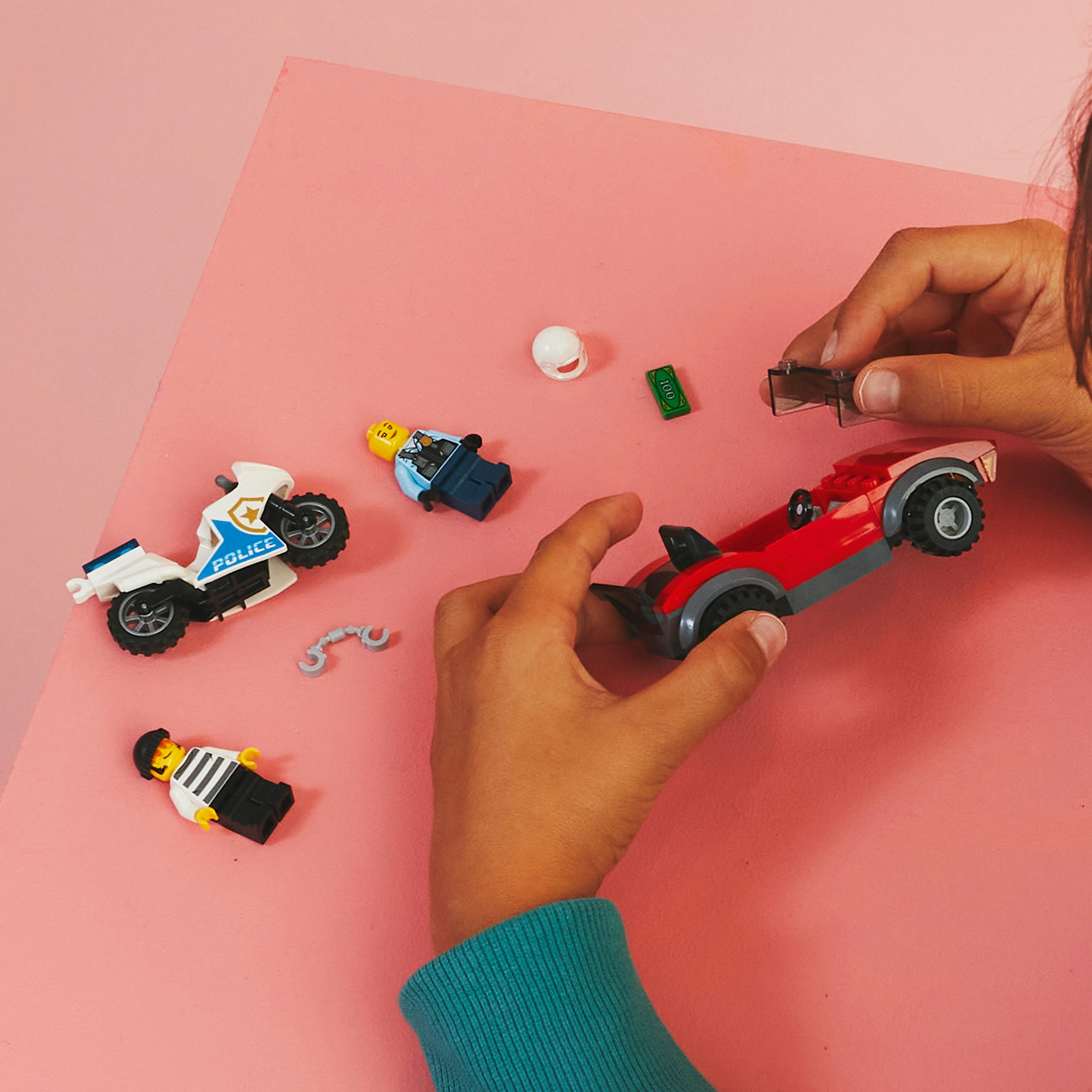 Lego speelfiguren en een rode auto zijn gerangschikt op een roze oppervlak. De handen van een kind zijn zichtbaar.