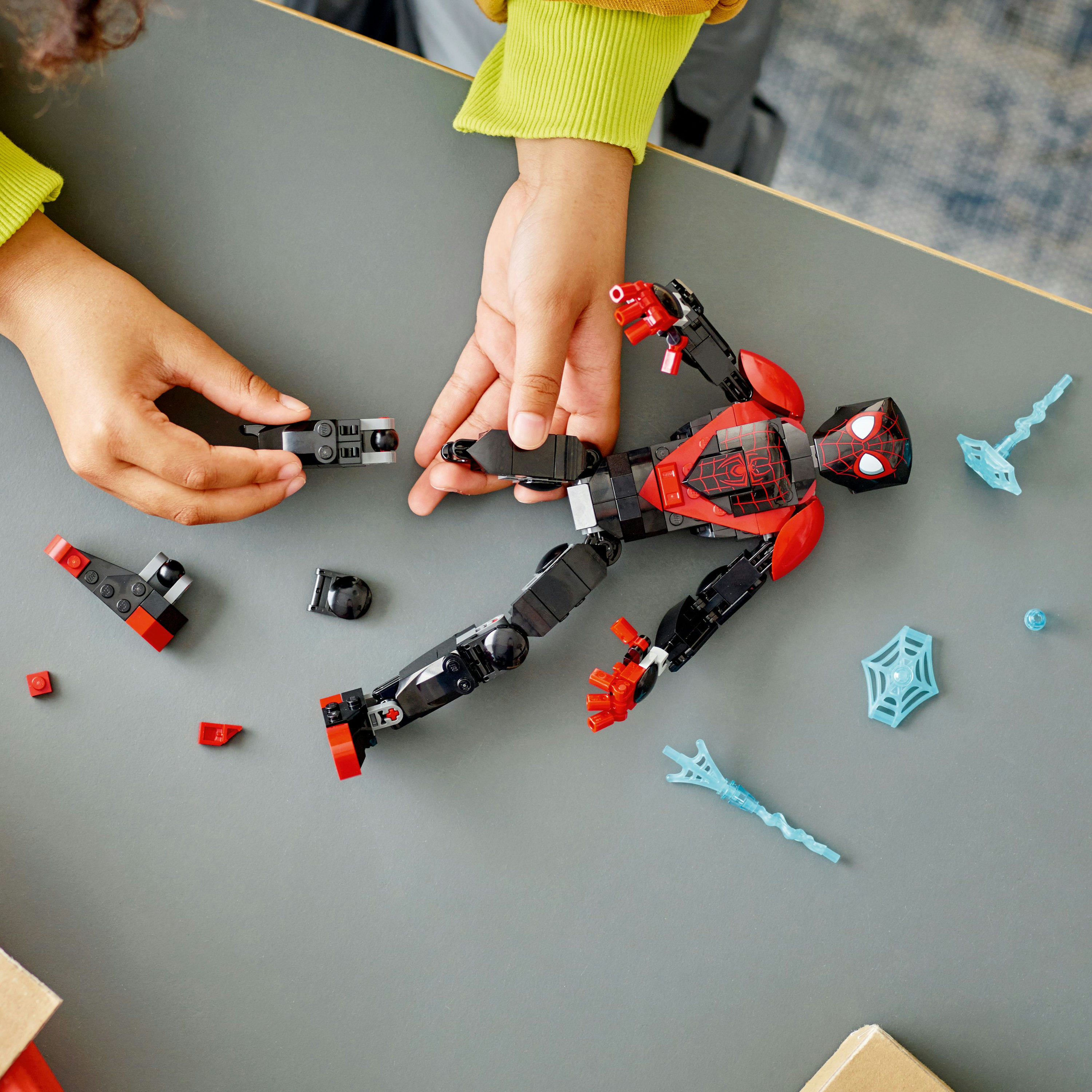 Hände, die eine rote und schwarze LEGO Spiderman-Figur auf grauer Oberfläche zusammensetzen.