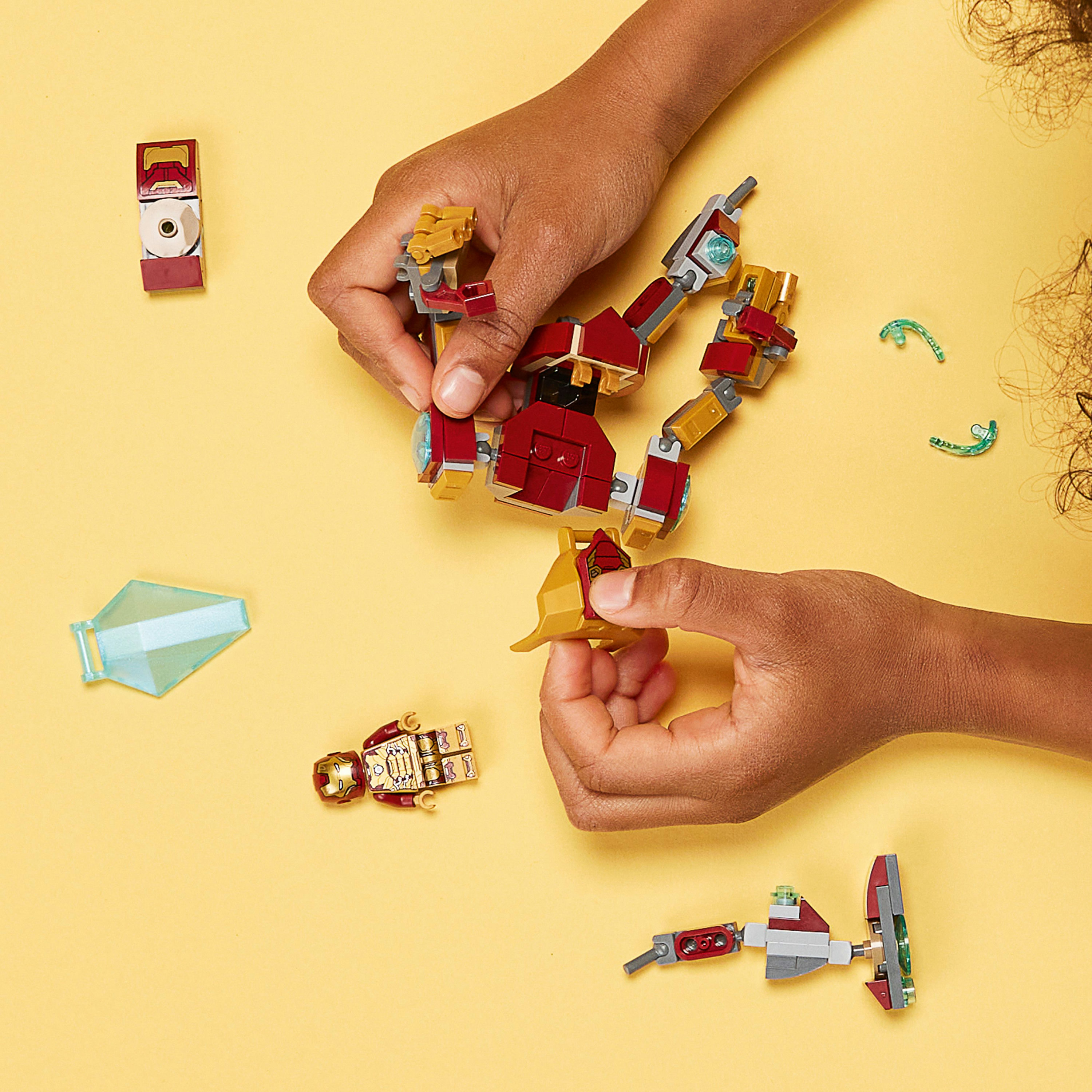 Mani che assemblano la figura Lego Iron Man. Pezzi Lego di vari colori su una superficie gialla.
