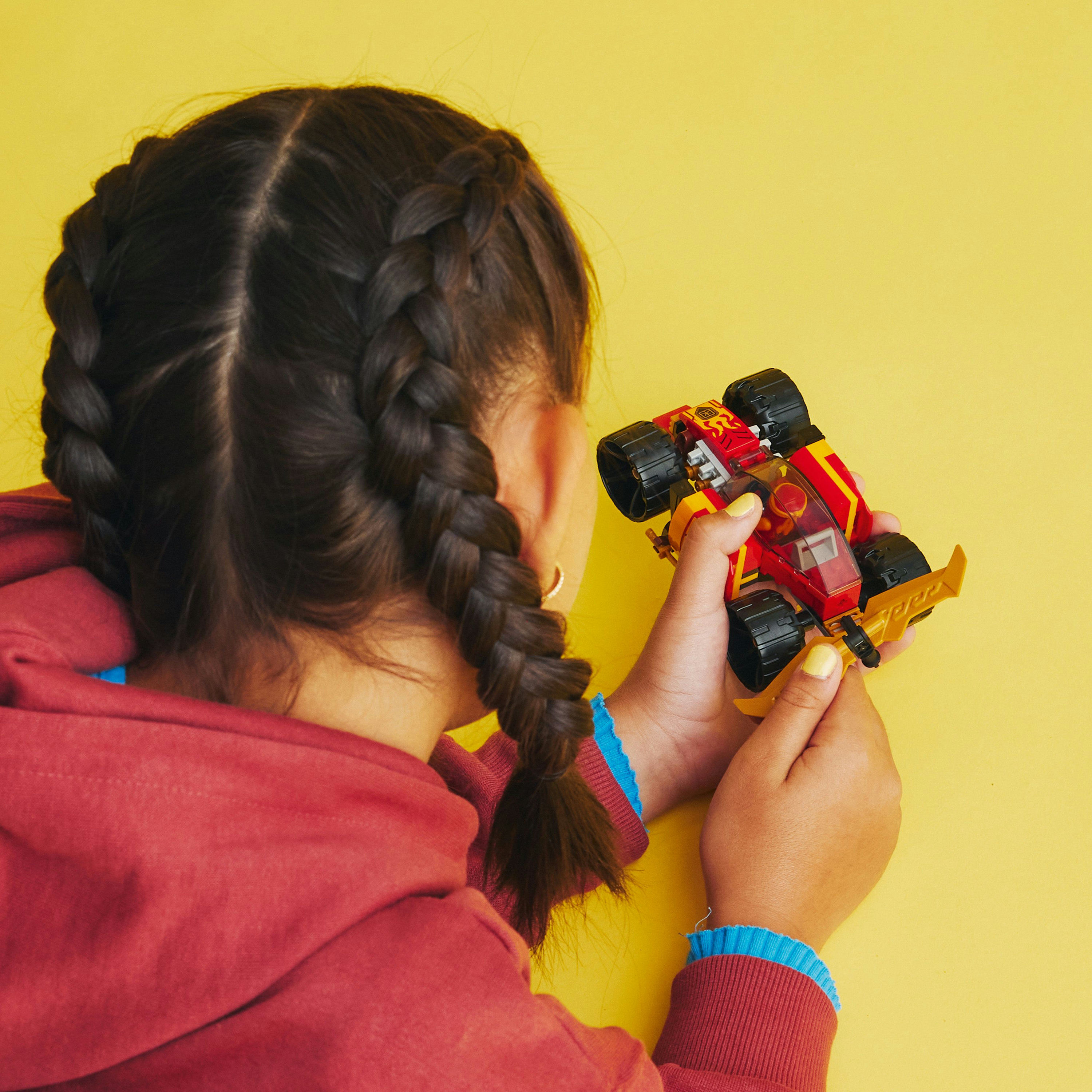 Dziecko trzymające czerwono-żółty samochód Lego. Ma warkocze i czerwoną koszulę.