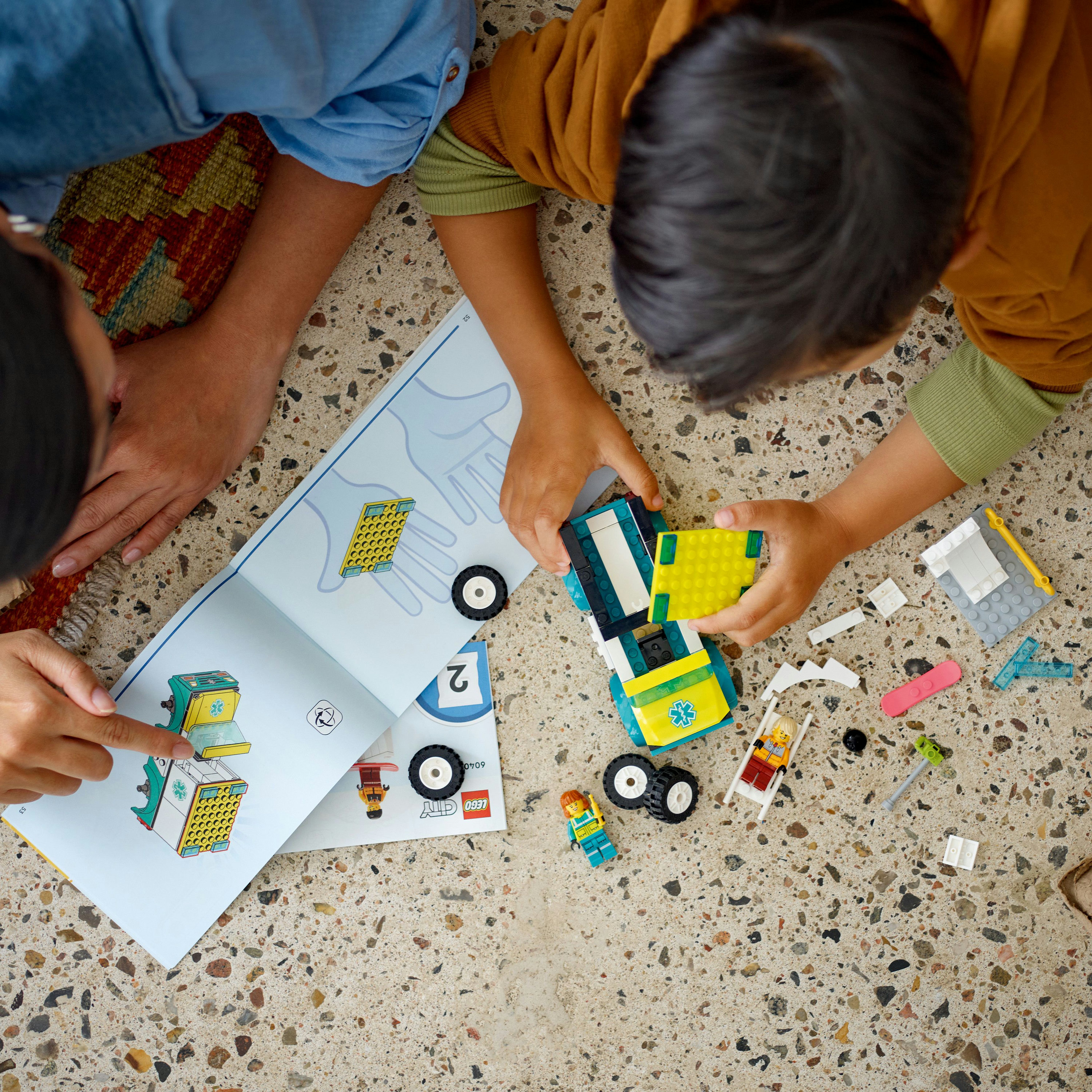 İki kişi birlikte Lego yapıyor, talimatlar ve Lego parçaları bir zemine yayılmış.