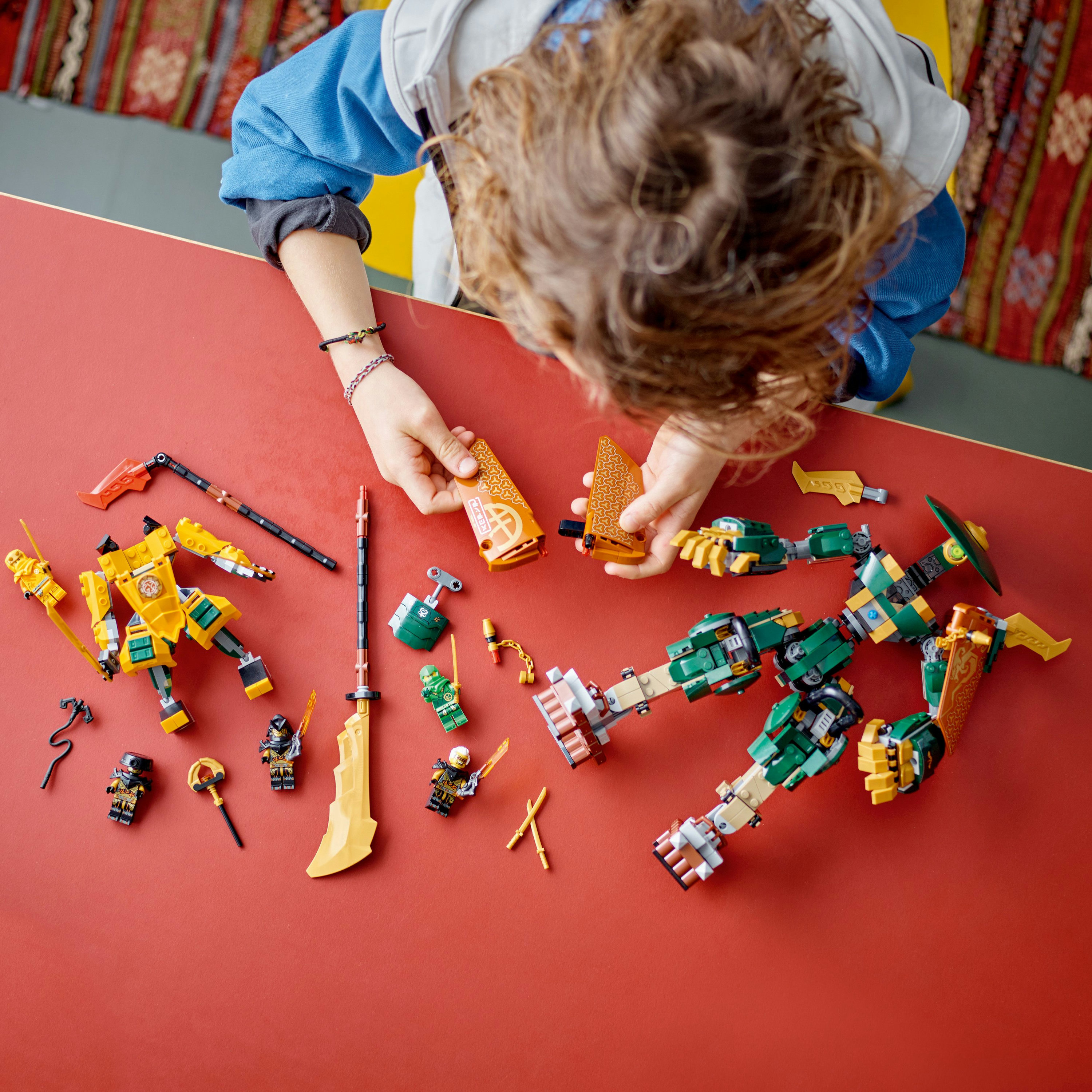 Een kind assembleert Lego Ninjago figuren op een rode tafel. Diverse Lego onderdelen zijn verspreid.