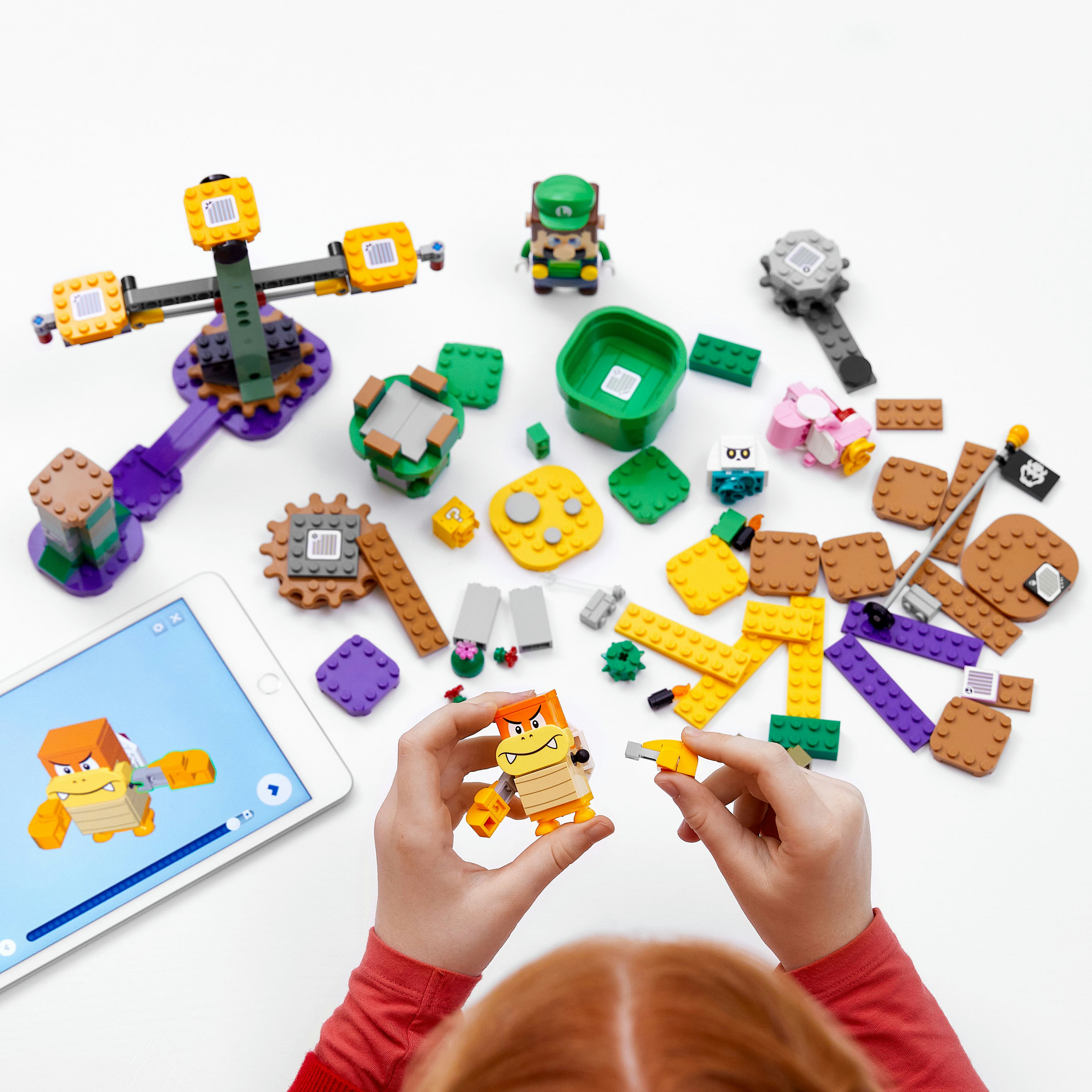 Kinderhände, die LEGO Mario-Set-Teile zusammenbauen, mit anderen Figuren und einem Tablet, das Anweisungen zeigt.