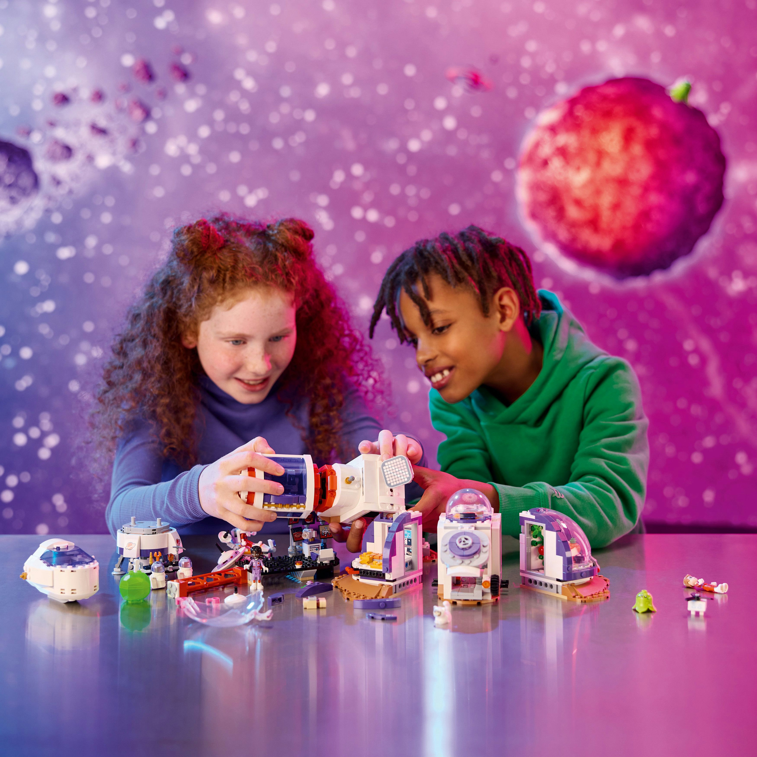 Twee kinderen bouwen een LEGO ruimtestationset. Diverse LEGO-onderdelen en modellen op een tafel.