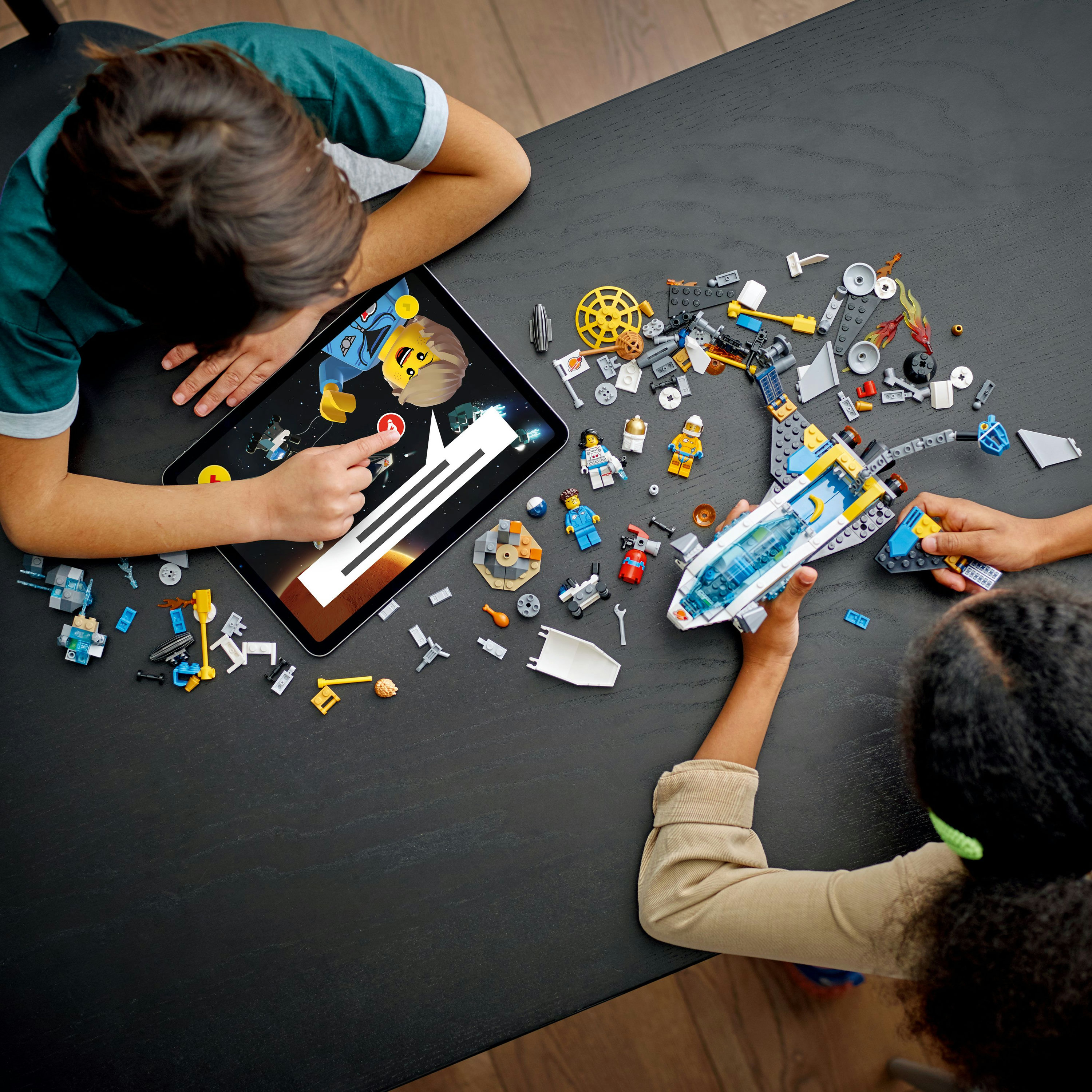 Zwei Kinder bauen ein Lego-Raumschiff. Eines benutzt ein Tablet. Viele Legoteile liegen auf dem Tisch.