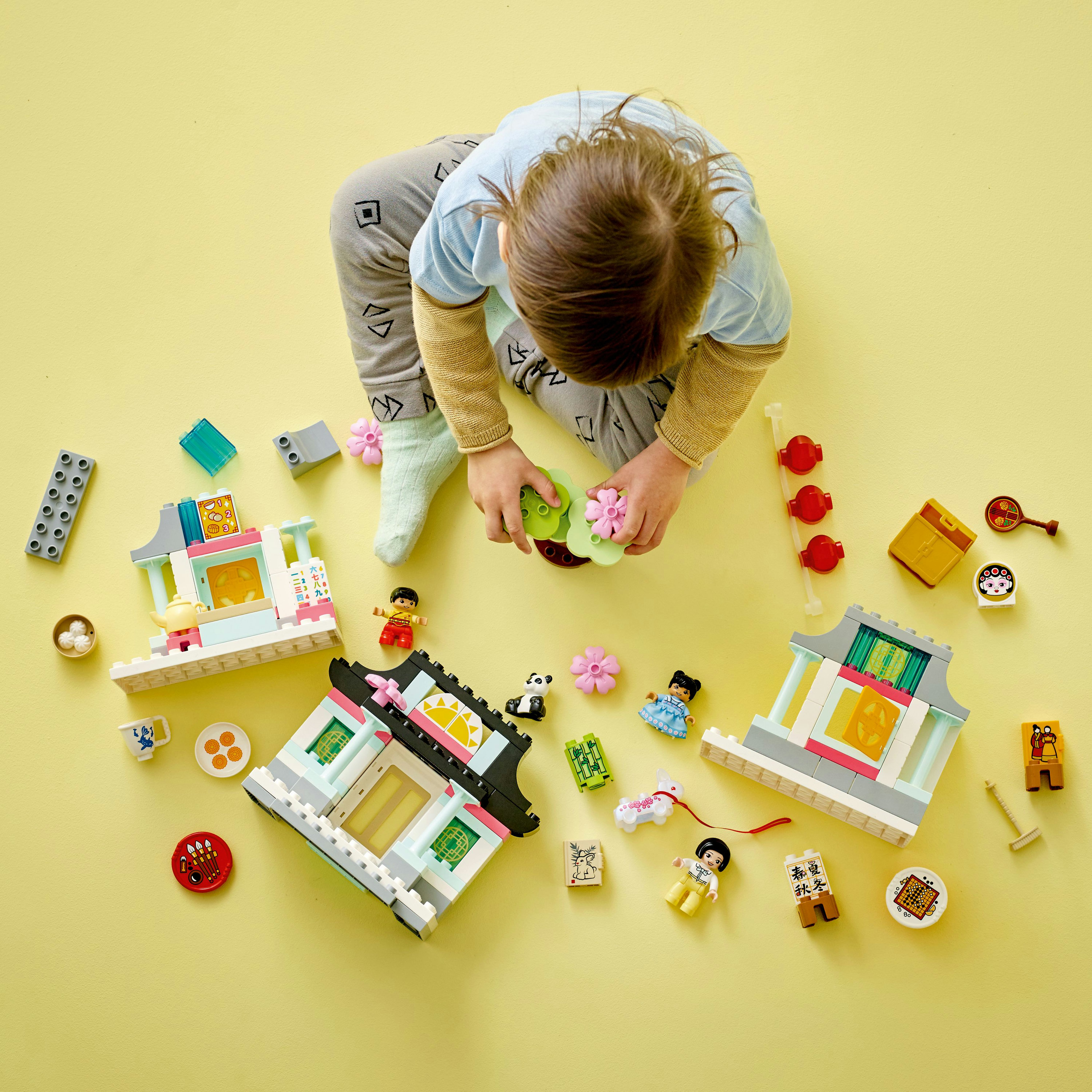 Un bambino gioca con un set LEGO con edifici e minifigure su una superficie gialla.