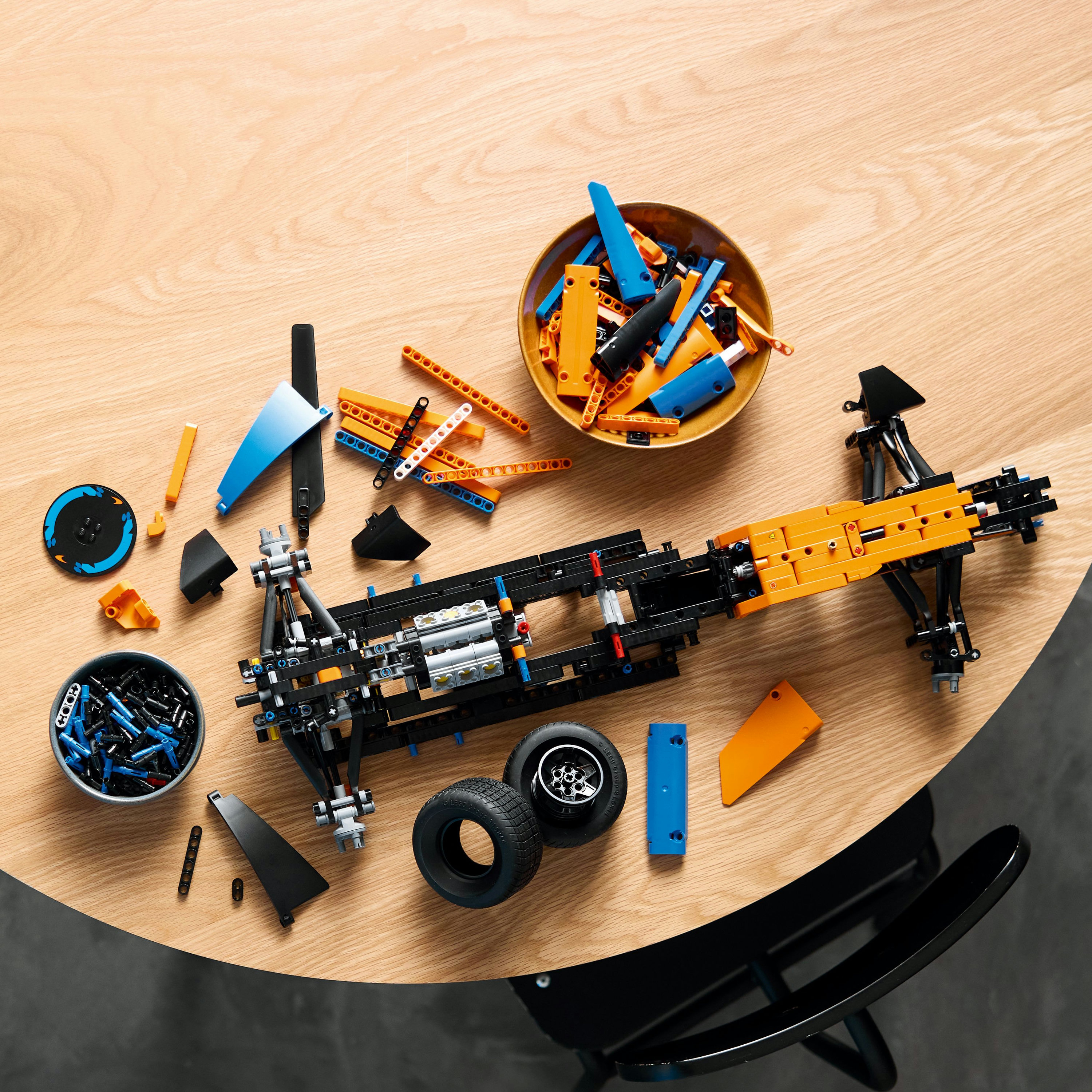 Części Lego Technic rozrzucone na drewnianym stole z czarnym krzesłem.