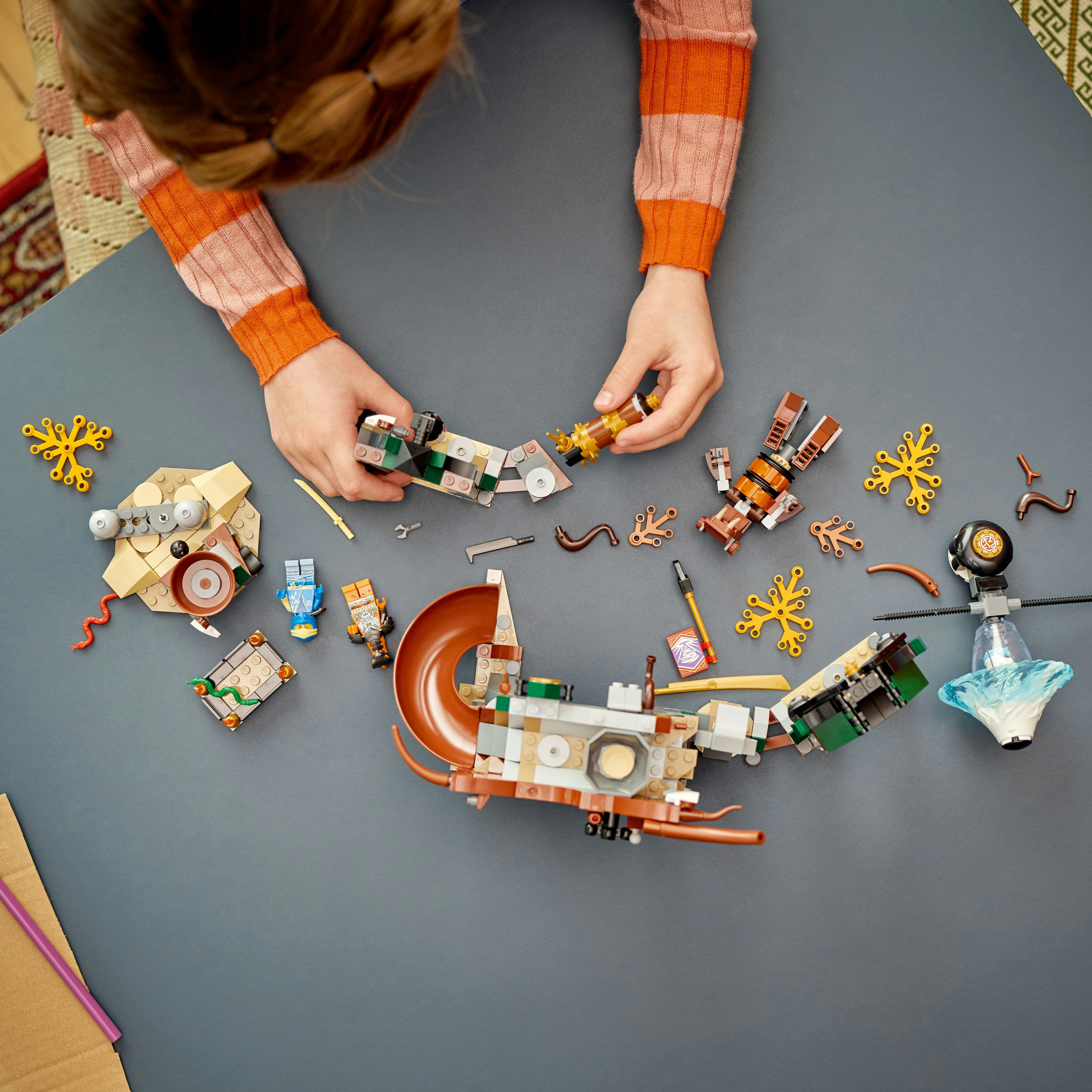 Hands assembling a LEGO set with small vehicle parts and other LEGO bricks on gray table.