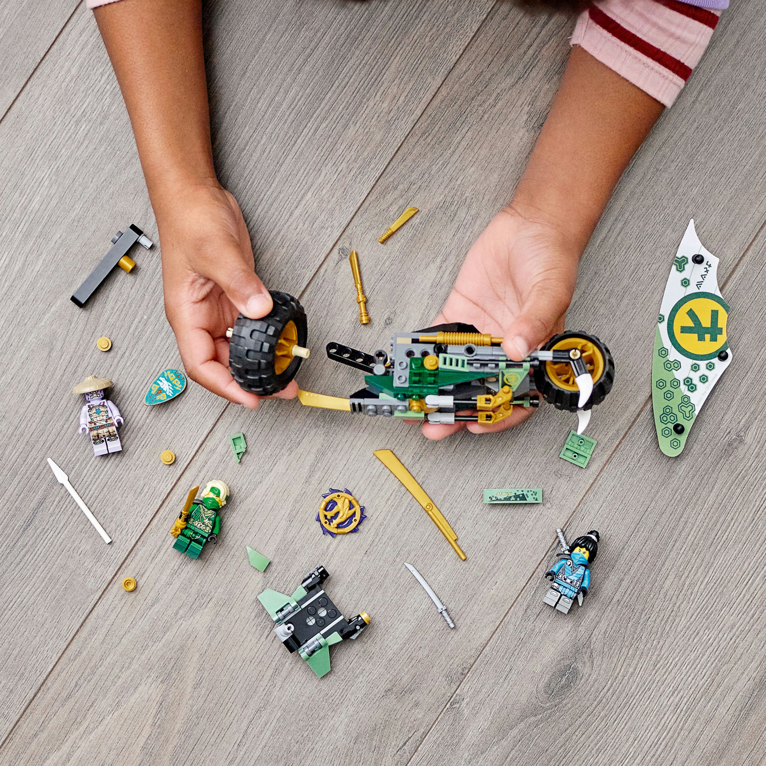 Hände, die ein Lego Ninjago Set mit Figuren, Waffen und einem Fahrzeug auf Holz zusammensetzen.