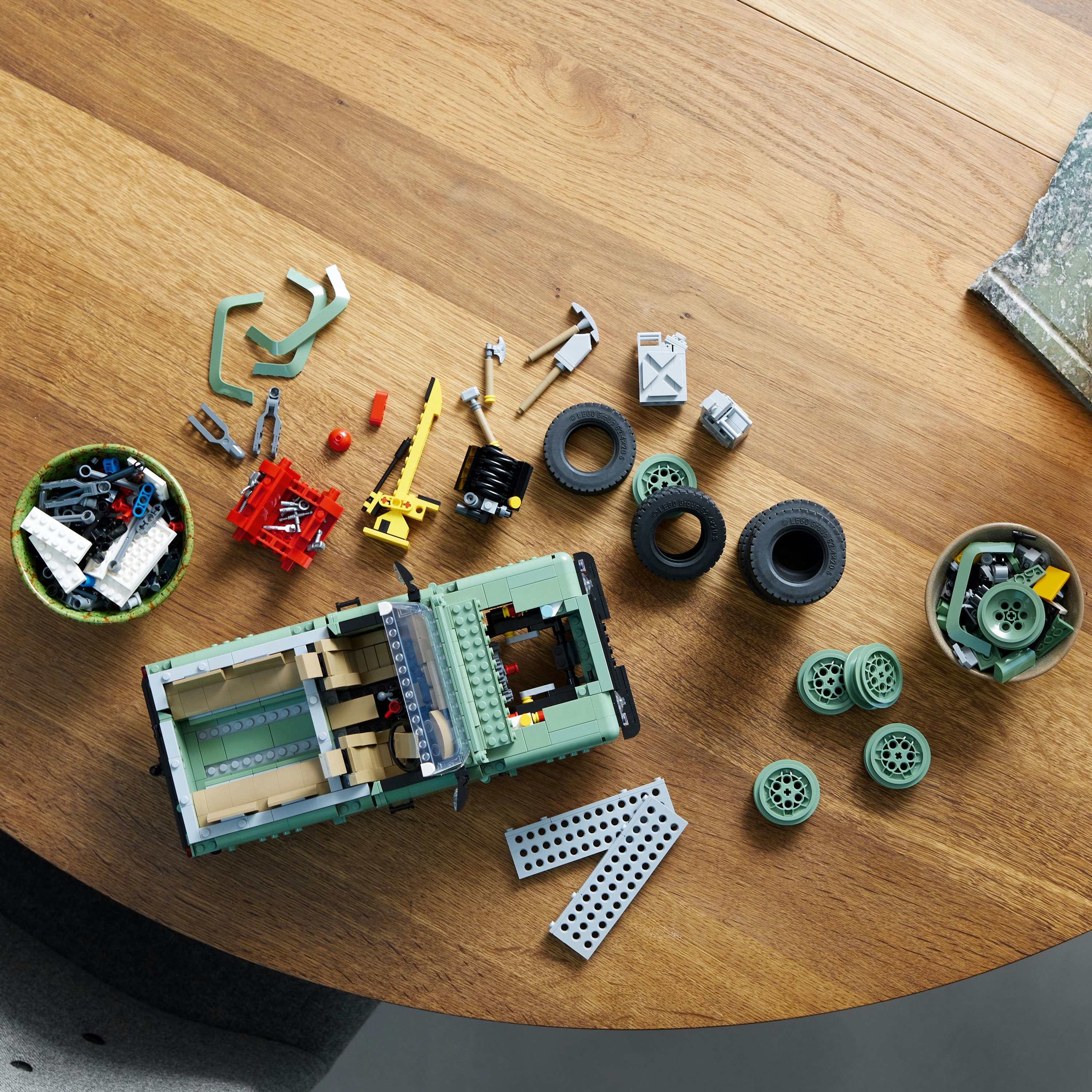 Drewniany stół z częściami samochodowymi Lego. Zbudowany jest zielony samochód. Są miski z narzędziami.