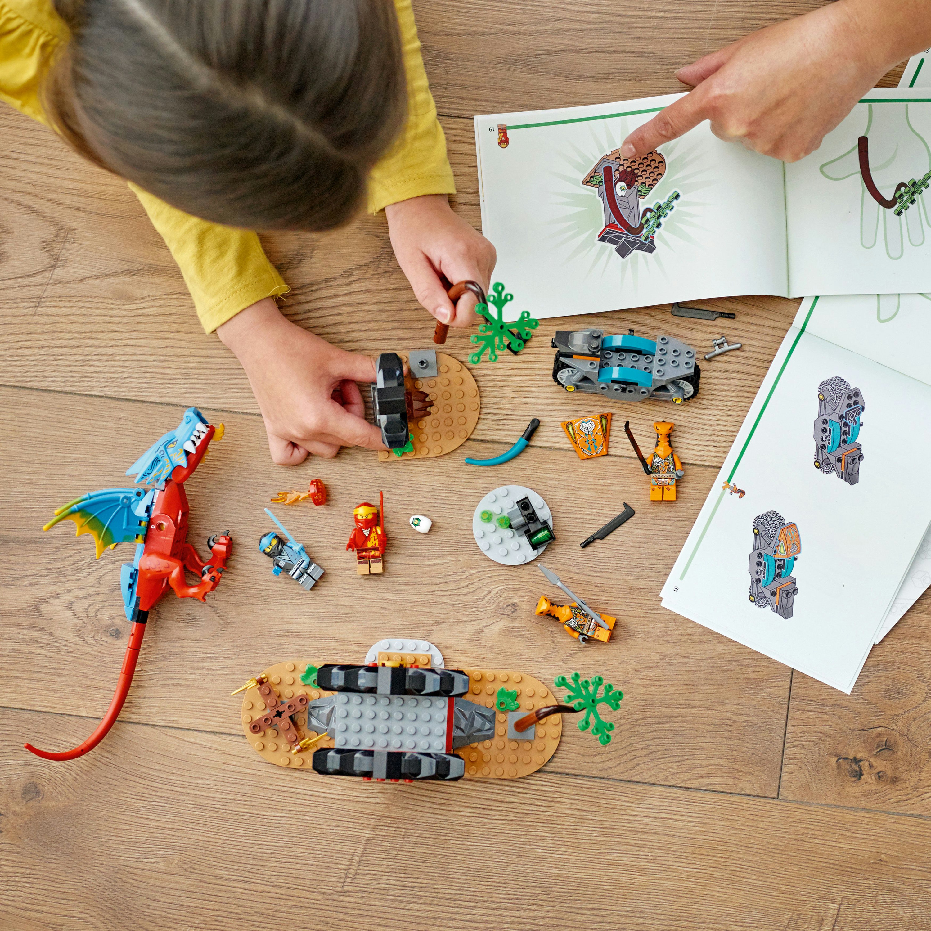Kinderhände, die LEGO-Teile zusammensetzen, mit einem Drachen und anderen Spielzeugteilen auf einer sandigen Spielmatte verstreut.