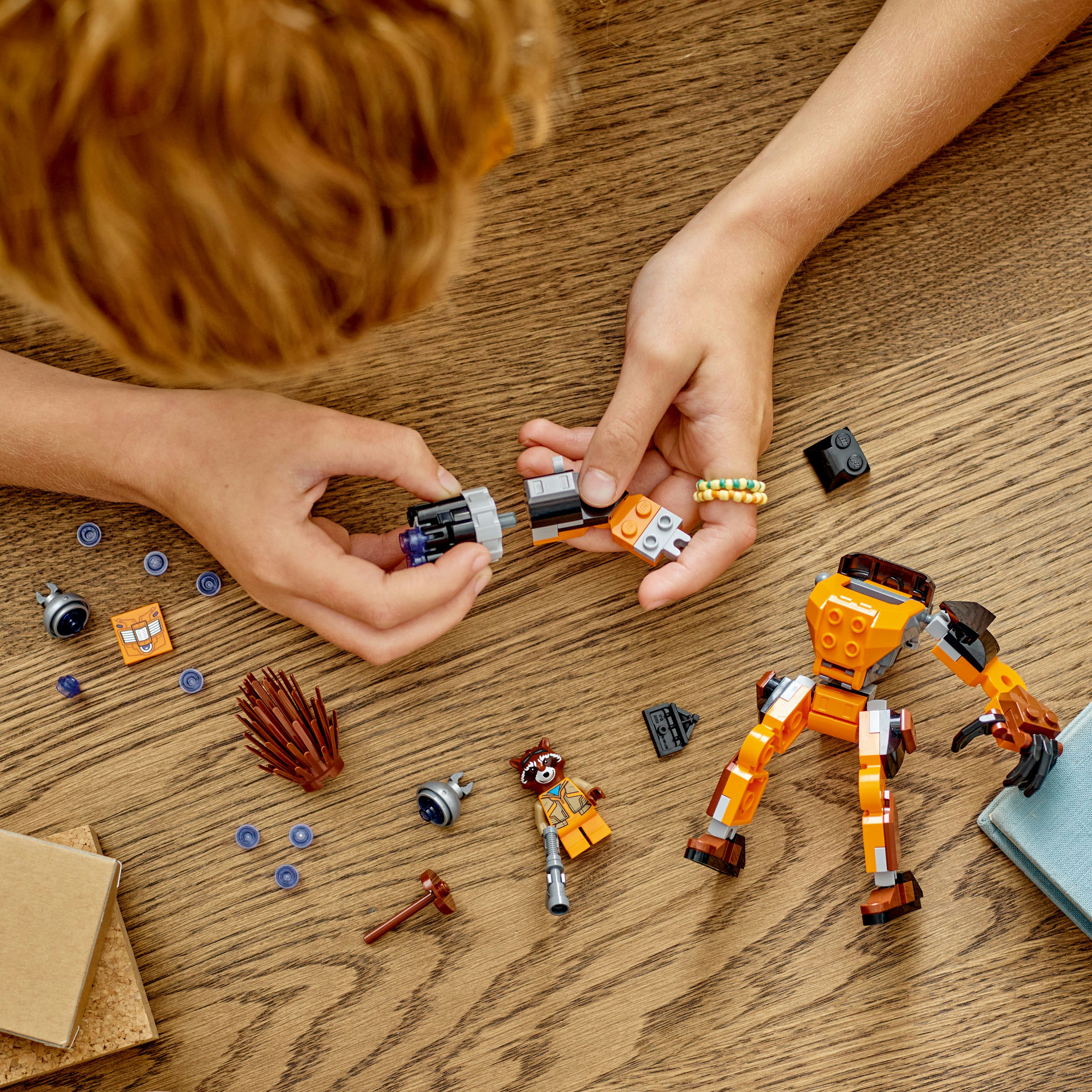 Handen die Lego-stukken in elkaar zetten. Oranje en grijze robotonderdelen, blauwe cirkelstukken en een figuur liggen op een tafel.