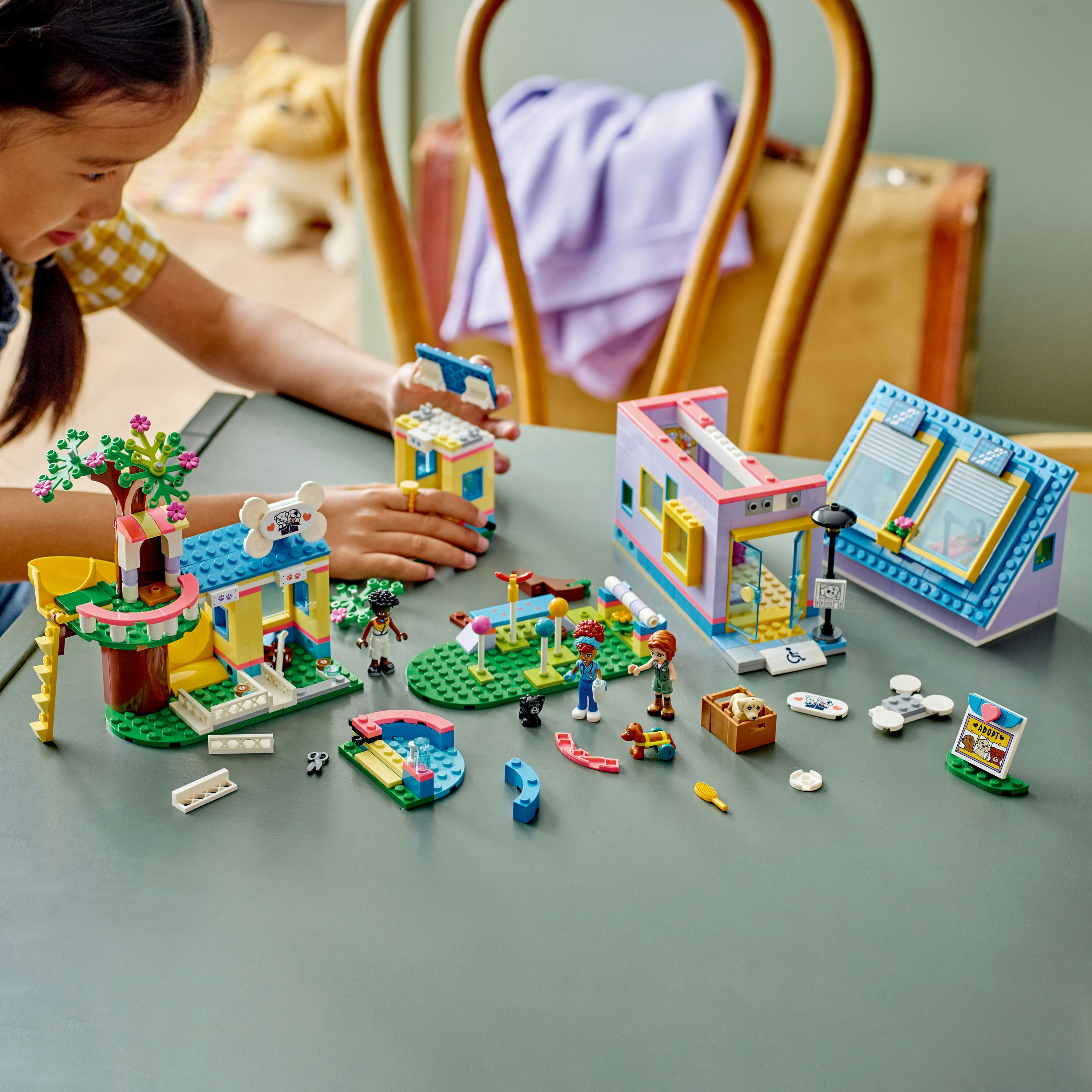 Kız Lego Friends setiyle oynuyor. Gri masa üzerinde renkli binalar ve figürler. Arka planda köpek oyuncağı.