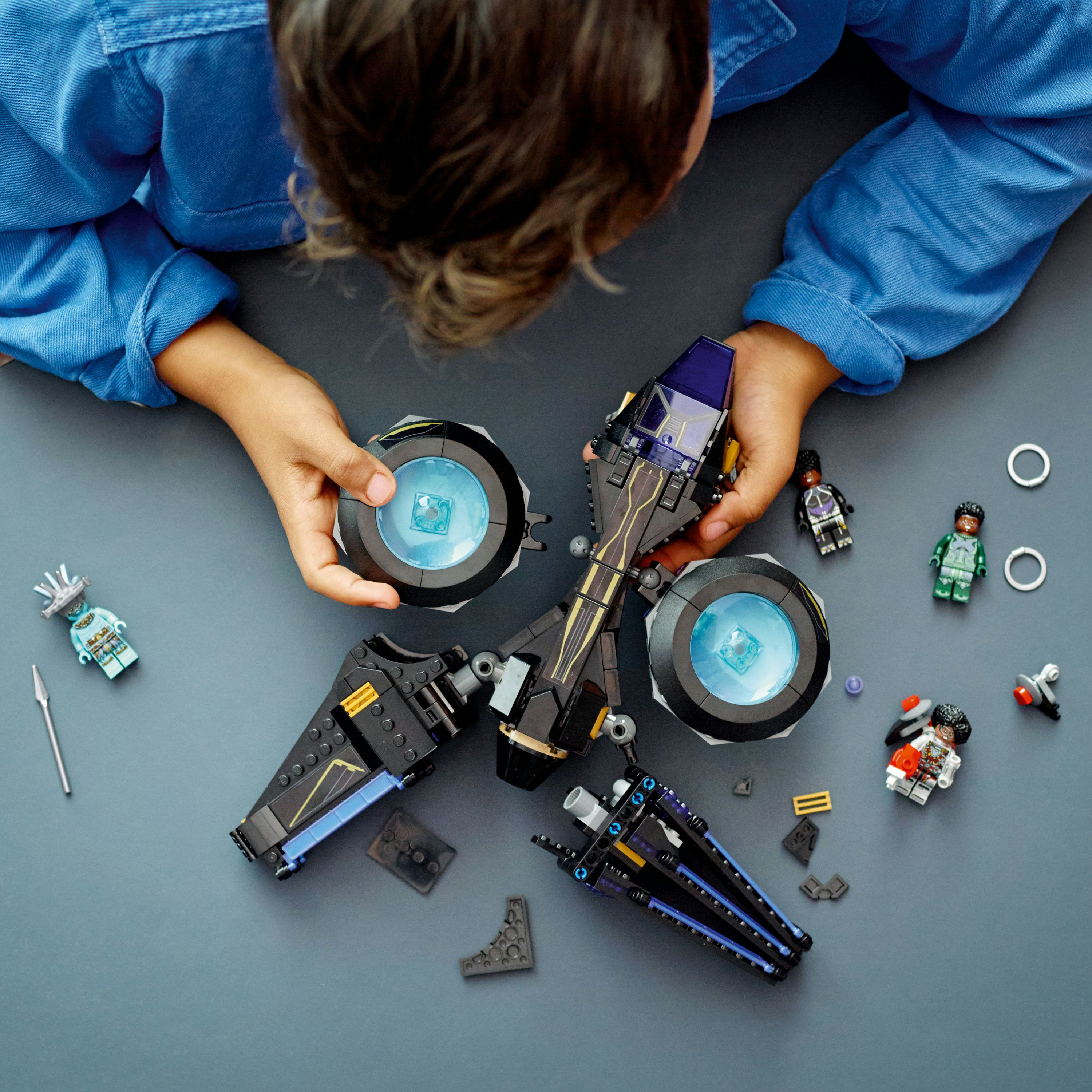 Dziecko składające czarno-niebieski pojazd Lego z figurkami na szarej powierzchni.