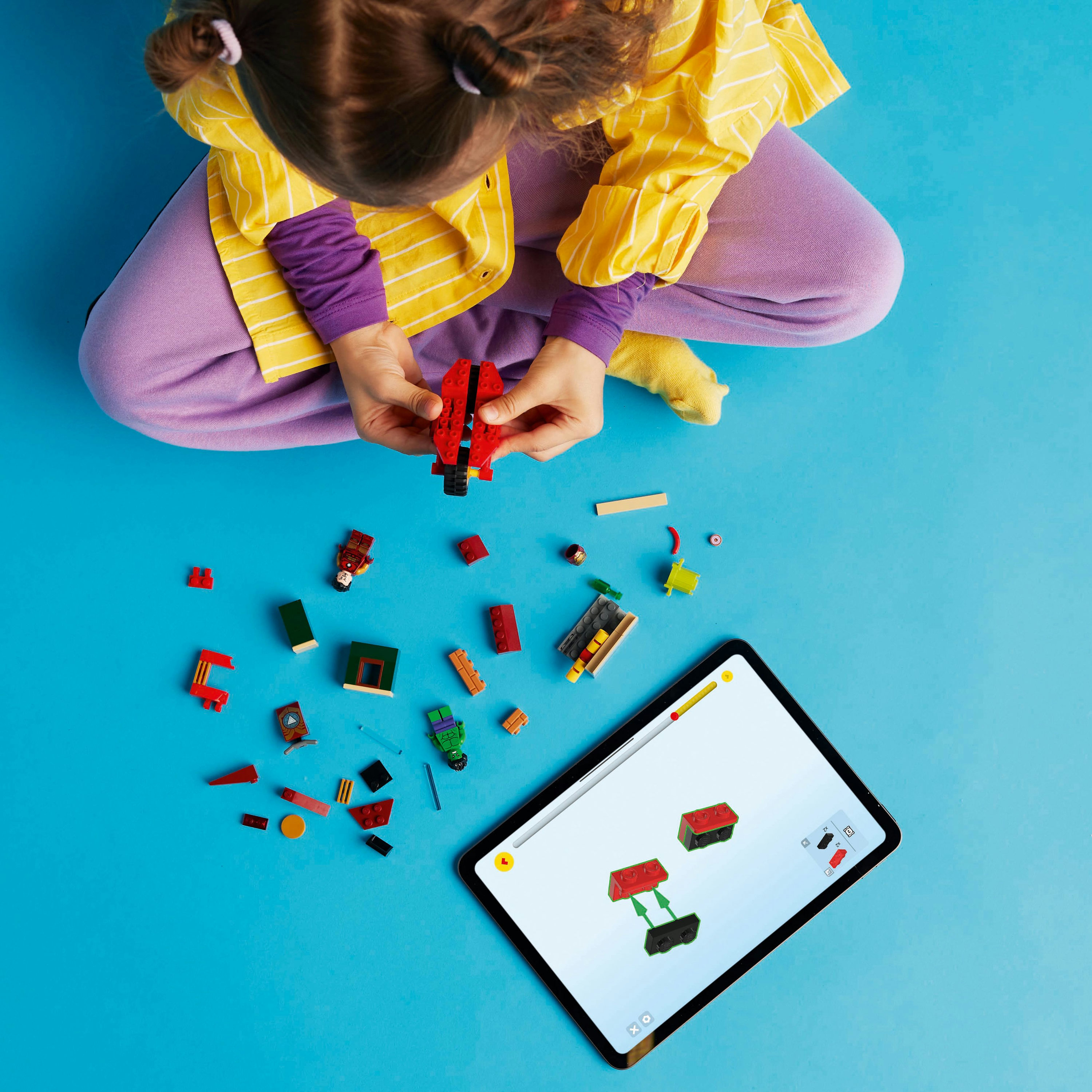 Dziecko z klockami Lego i tabletem. Klocki rozrzucone na niebieskiej powierzchni.