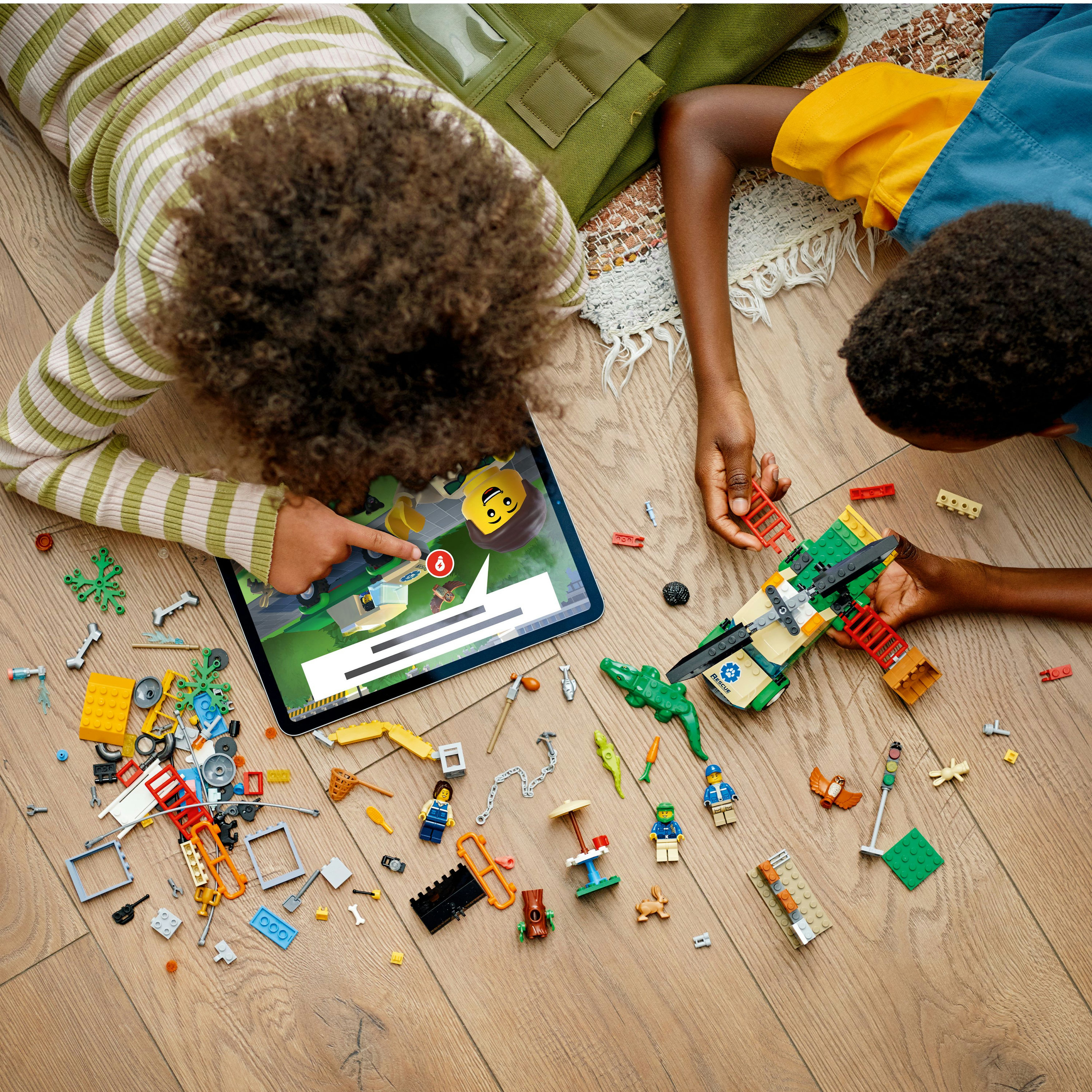 Zwei Kinder spielen auf einem Holzboden mit LEGOs. Ein Tablet zeigt ein Spiel. LEGO Steine sind verstreut.