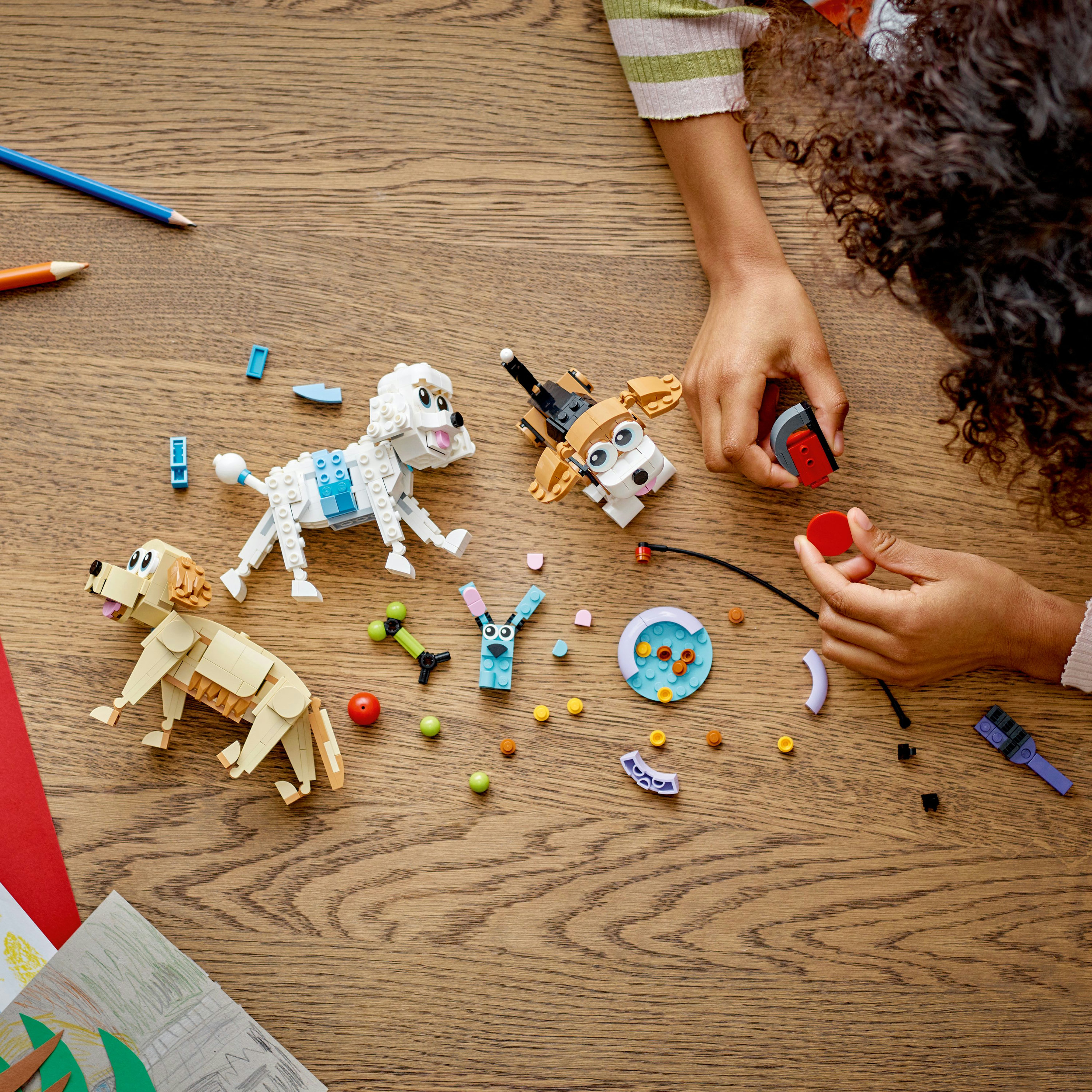 Handen van een kind dat speelt met Lego-hondmodellen en andere Lego-accessoires op een houten tafel.