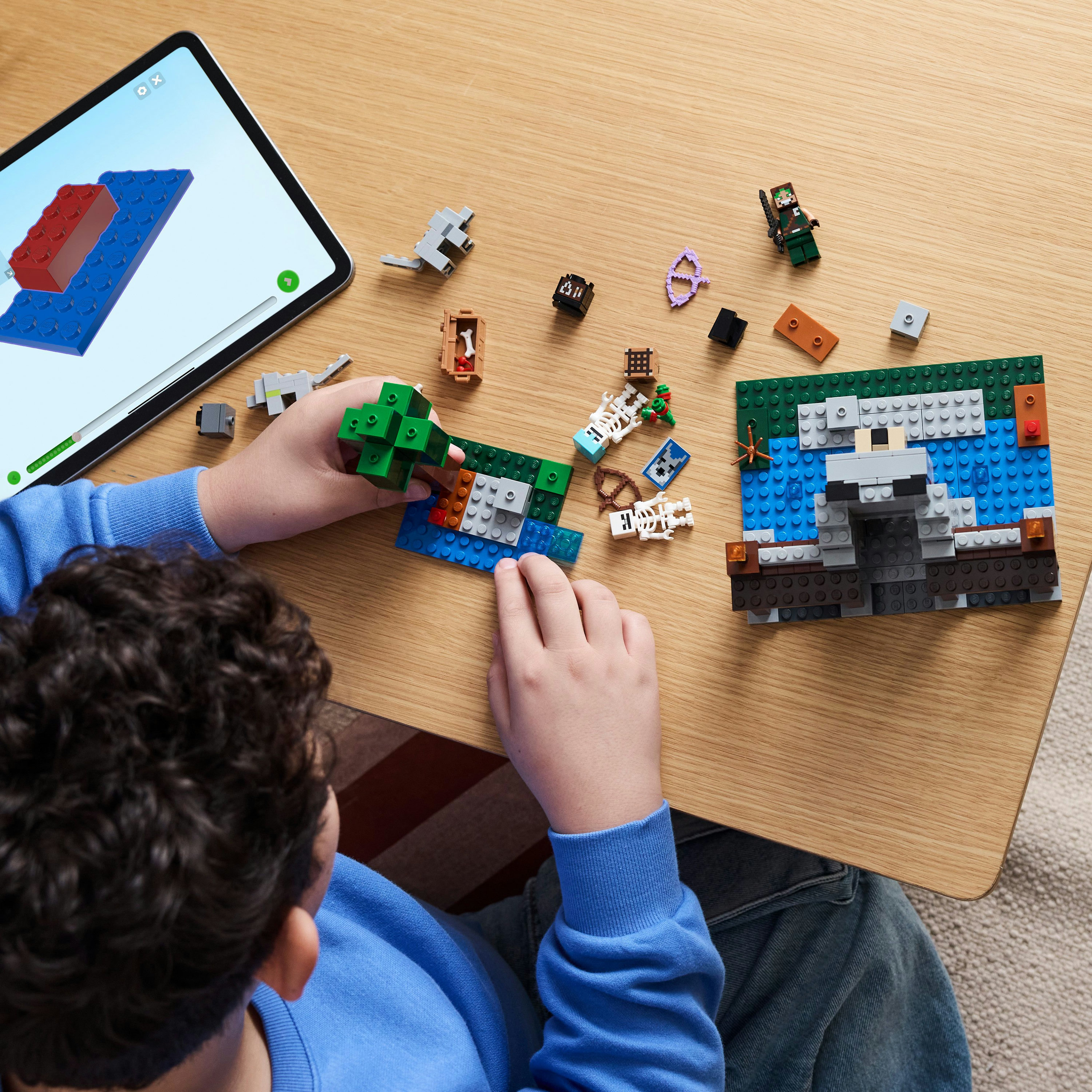 Ein Kind spielt mit einem Lego-Set auf einem Holztisch, mit einem Tablet, das Anweisungen zeigt. Draufsicht.