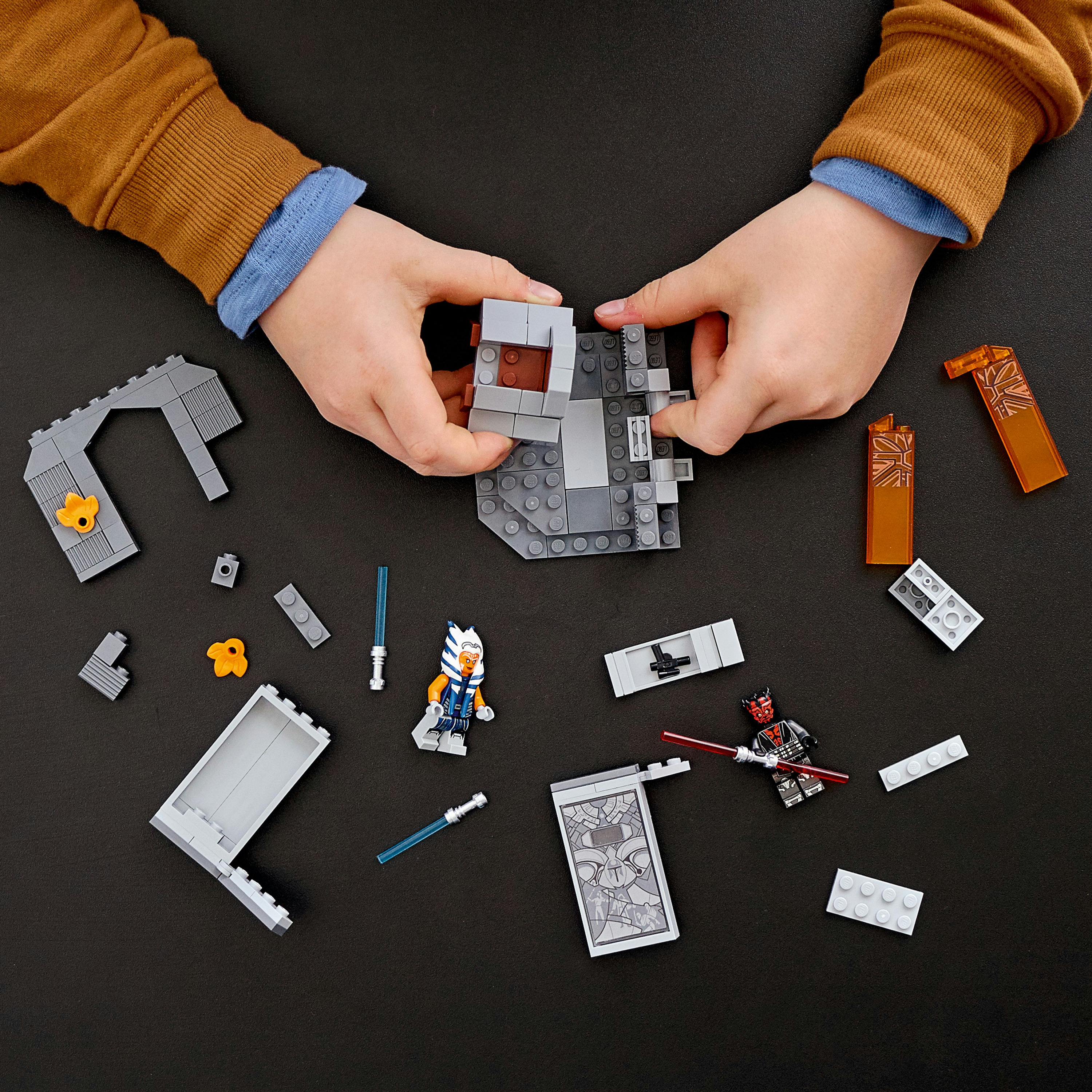 Hände bauen eine Lego-Struktur mit Minifiguren auf einer dunkelgrauen Oberfläche. Mehrere Teile.