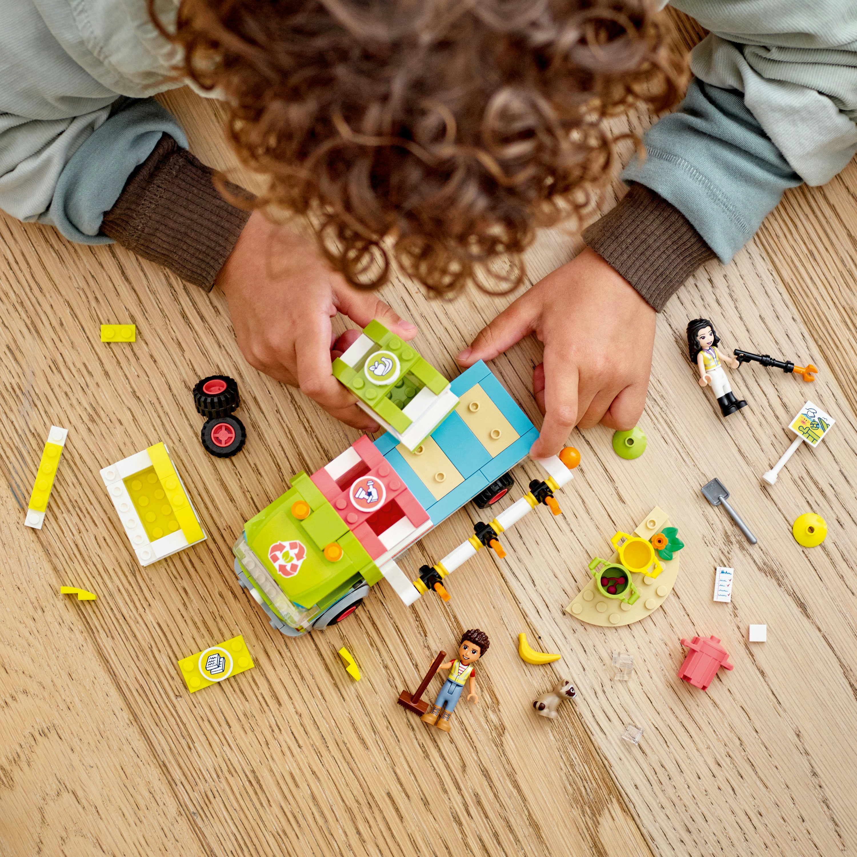 Kinderhände, die LEGO Friends Set auf einem Holzboden zusammensetzen, verschiedene Teile und Figuren verstreut herum.