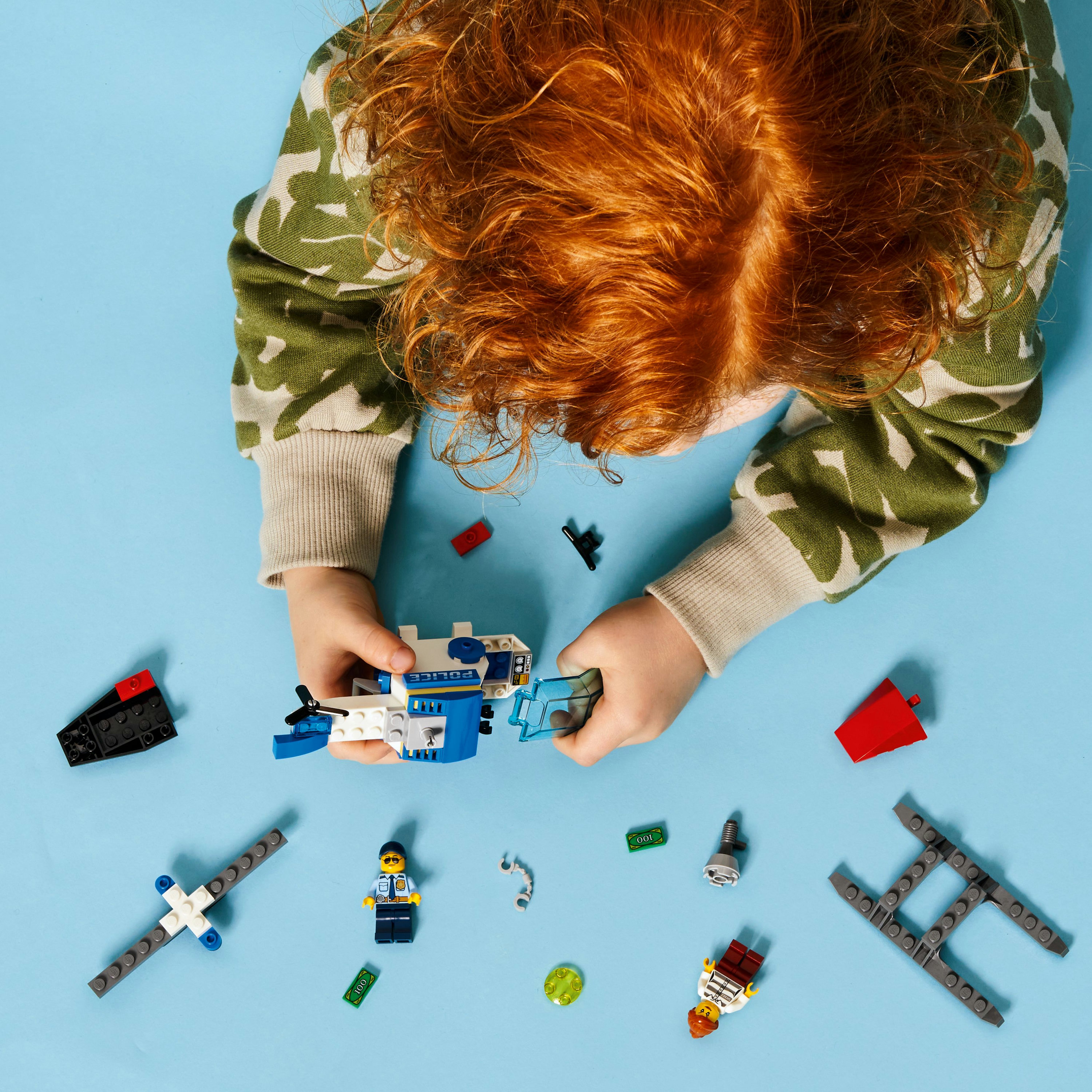 Ein Kind mit roten Haaren baut einen Lego-Hubschrauber auf einer blauen Oberfläche, mit verstreuten Lego-Teilen.