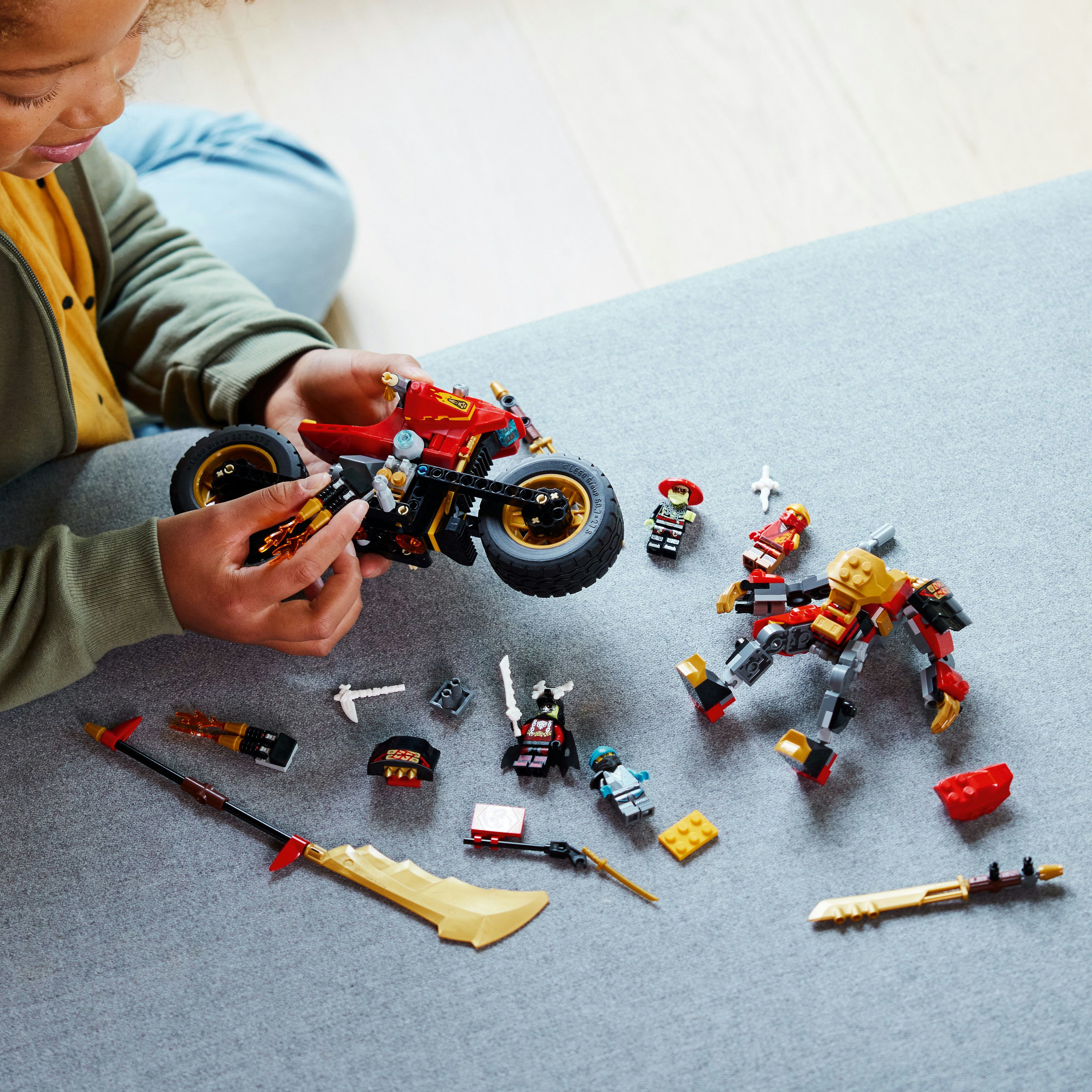 Een kind speelt met een Lego Ninjago set. Figuren en een motorfiets liggen op een grijs oppervlak.