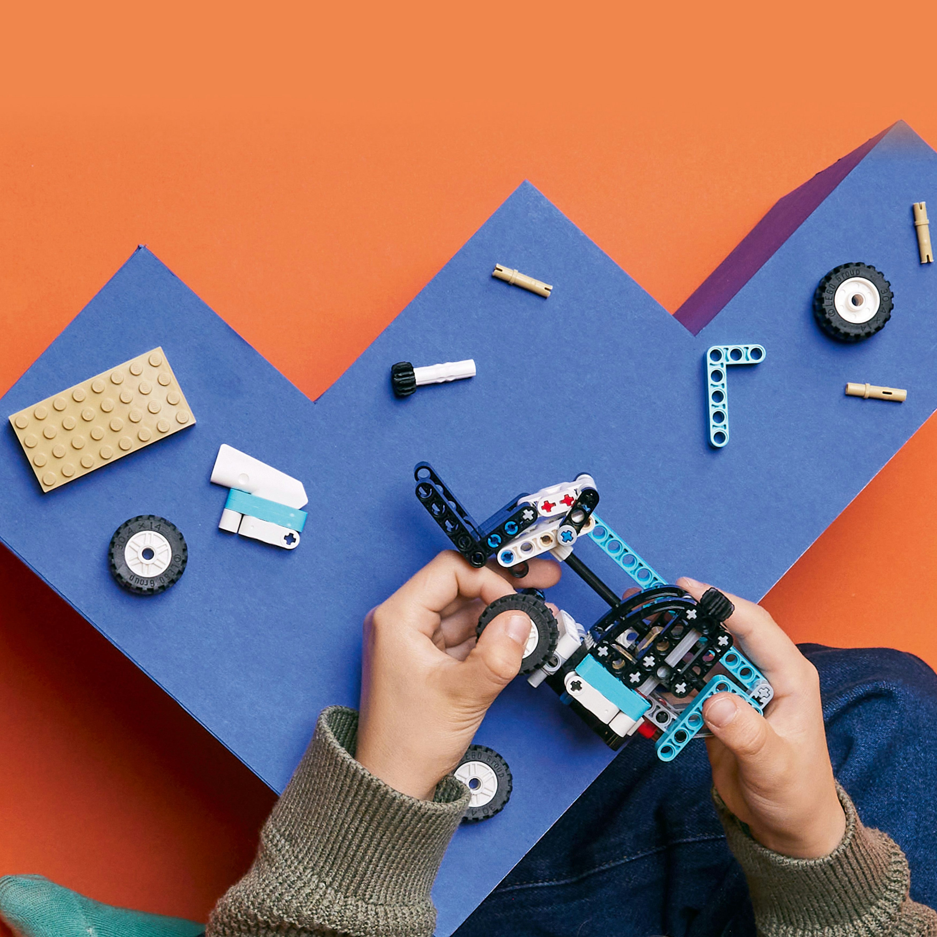 Hände bauen ein LEGO Technic Modell auf blauem und orangefarbenem Hintergrund. Verschiedene LEGO-Teile sind sichtbar.