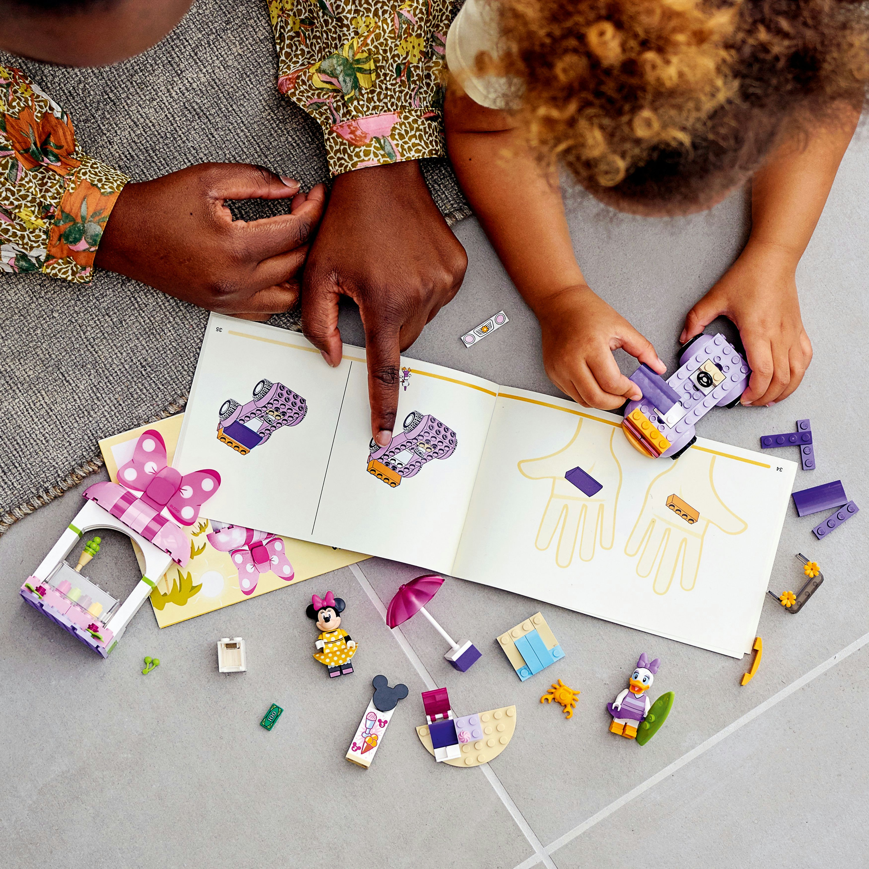 A person points to a Lego instruction manual. Lego toys are scattered on the floor.