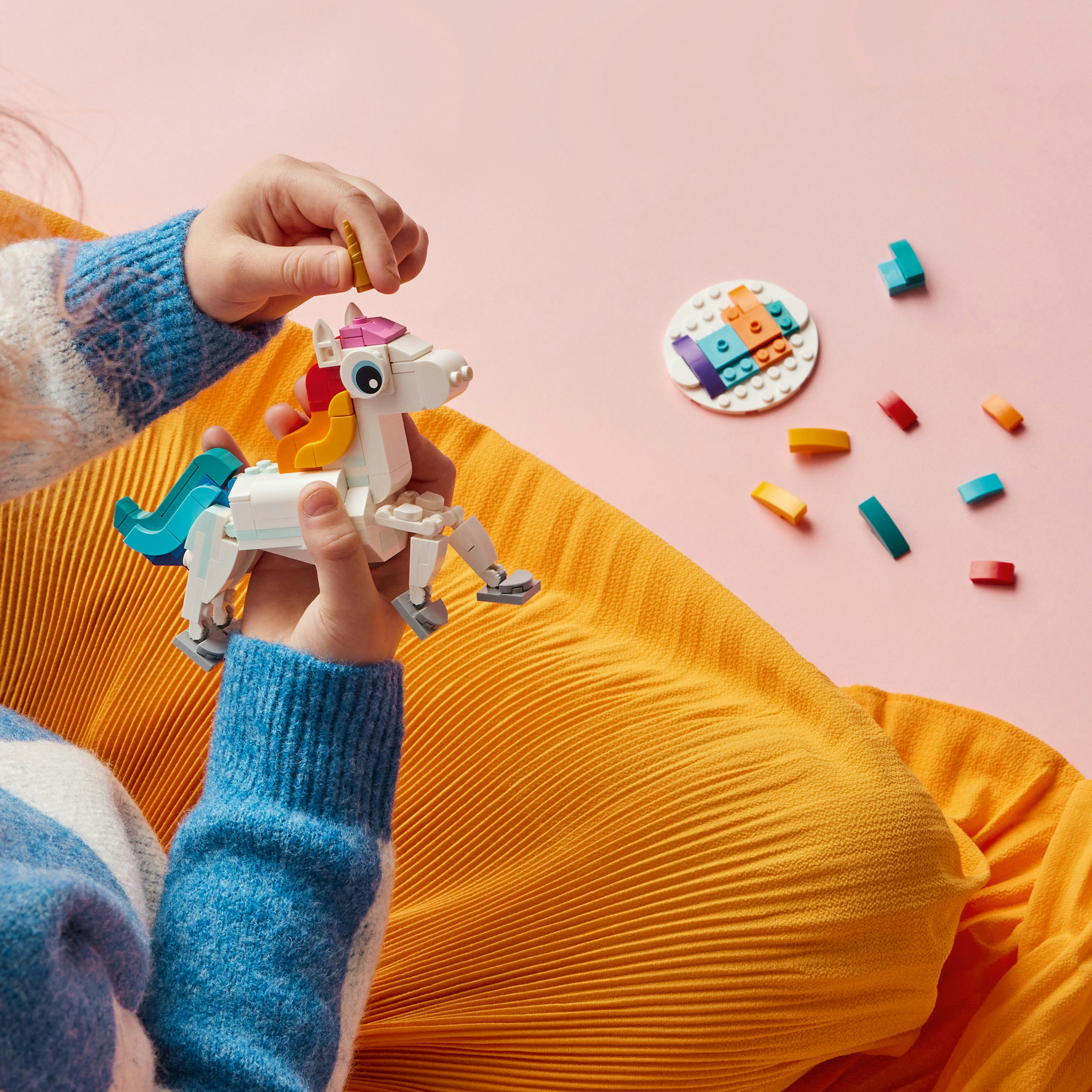 Osoba montuje białego jednorożca Lego z tęczowymi i niebieskimi detalami, na różowo-żółtej powierzchni.